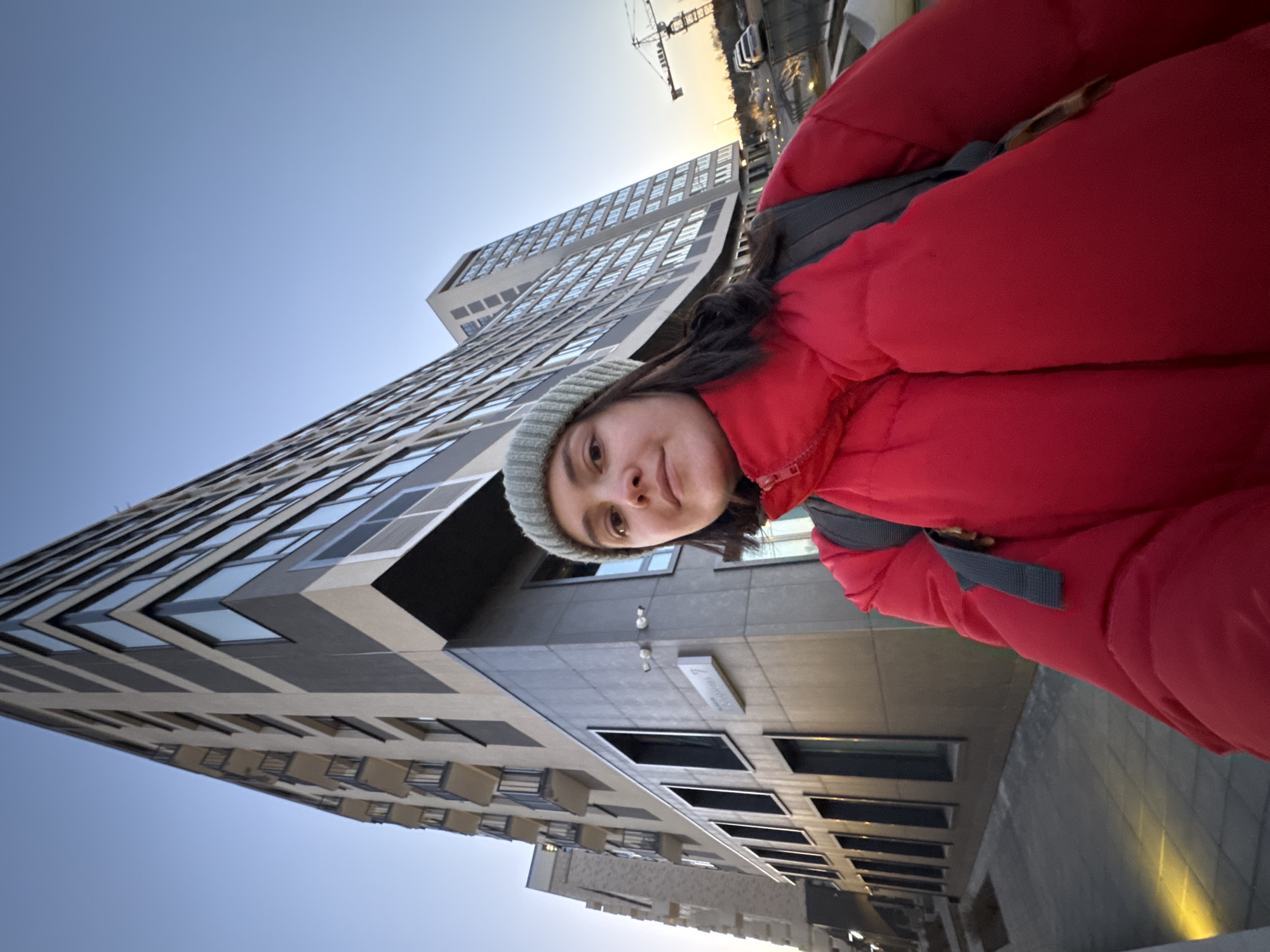 A woman in a red winter jacket and a light-colored beanie is positioned in front of a tall, modern building. The building has a grid-like pattern of windows and is made of light-colored materials. The sky is a clear blue, transitioning to a lighter hue near the horizon, indicating either sunrise or sunset. A construction crane is visible in the distance. The location is Izhevsk, Russia, based on the architectural style and building materials consistent with developments in the city.