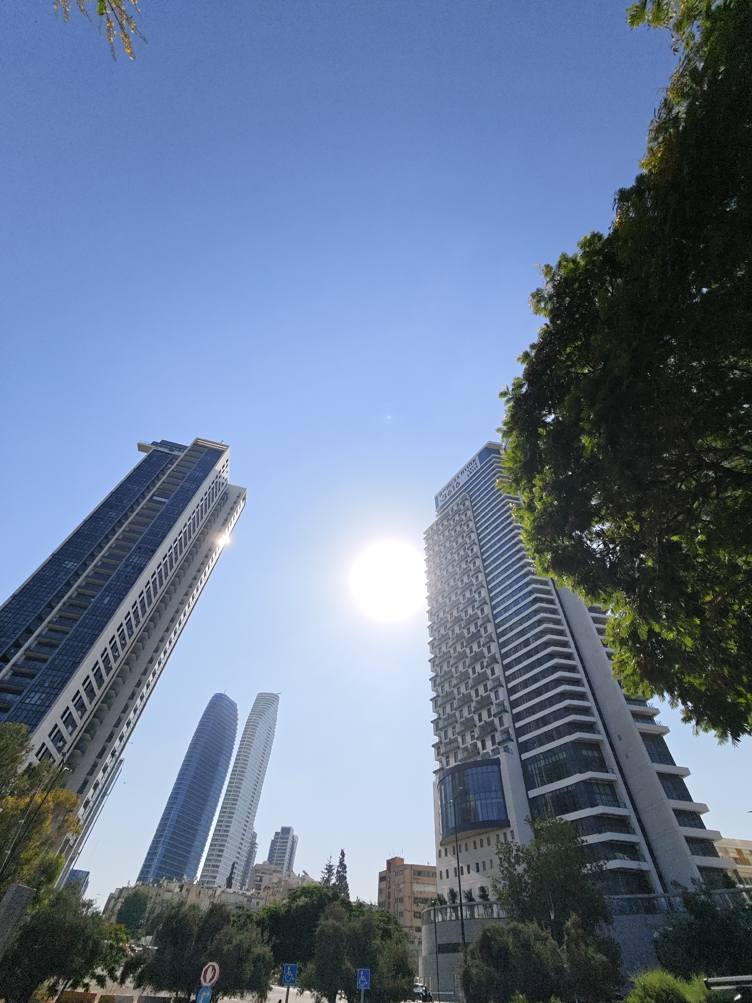 The image shows a street scene in Tel-Aviv, Israel, with several modern high-rise buildings. The sun is bright and positioned high in a clear blue sky, creating lens flare that obscures parts of the buildings. In the foreground, a large tree with green leaves partially obstructs the view of the right side of the scene.
Dominating the left side of the frame is a tall, slender skyscraper with a facade of dark glass and white horizontal bands. To its right, closer to the center, are two distinctive, curved skyscrapers with reflective blue and white facades, appearing to lean towards each other. Further to the right, another tall building with a white, angular facade, featuring numerous balconies and glass windows, is prominently visible. The sun's glare is most intense near this building.
In the background, other buildings of varying heights and architectural styles are visible, suggesting a dense urban environment. Some lower buildings and trees are also present, lining the street. There are no people or vehicles clearly discernible in the image, and no direct interactions are depicted. The overall impression is of a sunny day in a contemporary city.