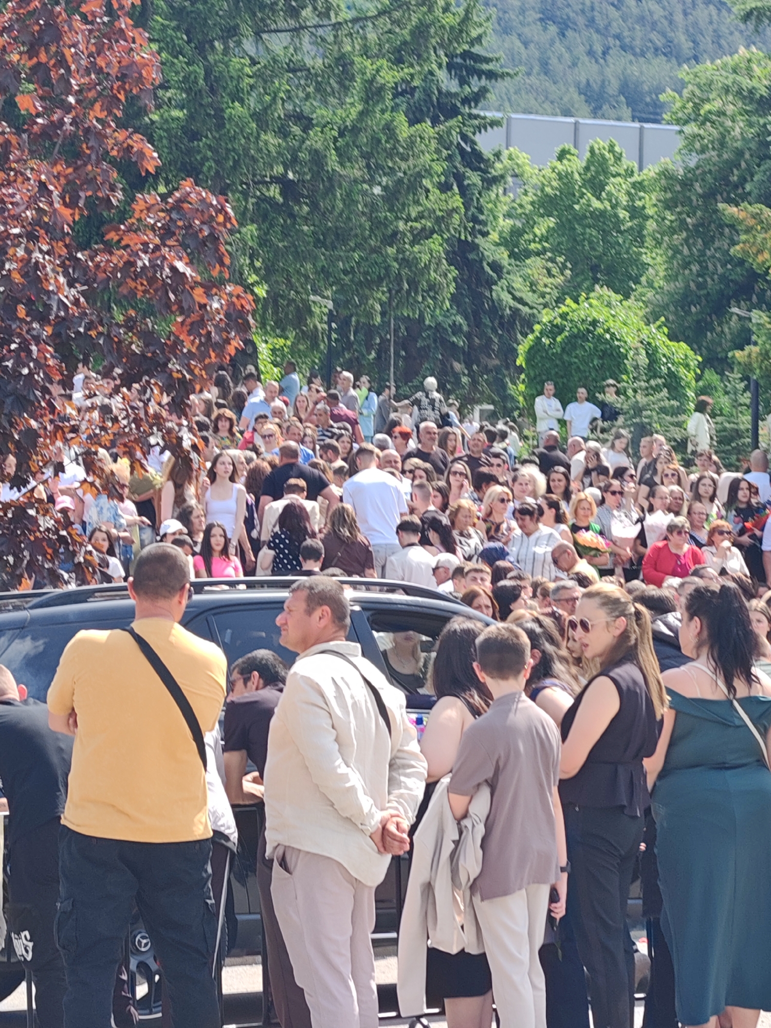 The image depicts a large crowd of people gathered in an outdoor setting. The location is Shumen, Bulgaria. The crowd is a mix of ages and genders, wearing a variety of clothing styles. A dark-colored car is visible in the foreground, partially obstructing the view of the crowd. Several individuals are standing near the car, seemingly observing the crowd. Behind the crowd are trees and possibly buildings. The scene appears to be a public event or gathering.