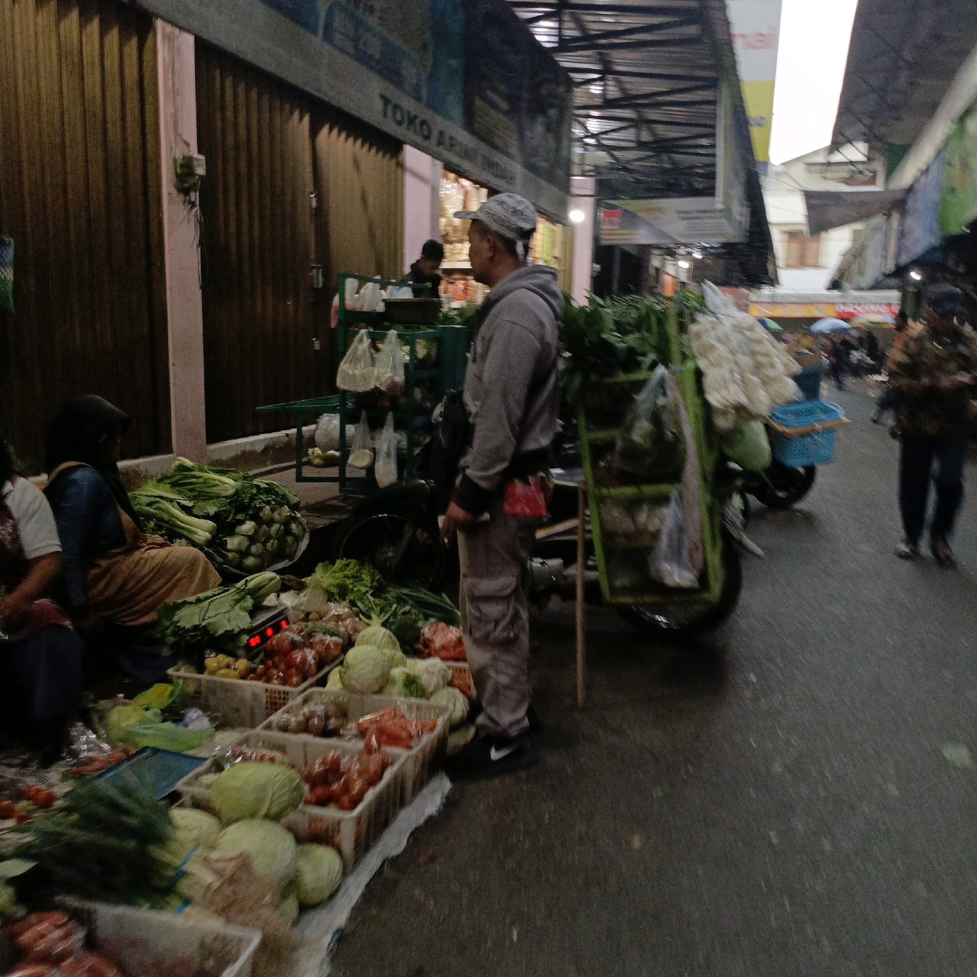 This is a street scene in Salatiga, Indonesia, featuring an outdoor market. In the foreground, there are numerous baskets filled with fresh produce, including cabbages, tomatoes, and leafy greens. A scale with a red digital display is visible among the vegetables.
To the left of the produce, a woman wearing a dark hijab and an orange skirt is seated, facing away from the camera. Another person, partially visible in the lower left, is wearing a white top and dark pants.
In the middle ground, a man wearing a grey hooded sweatshirt and light-colored pants stands facing right, looking at the market stalls. He has a patterned baseball cap on and a red cloth tied around his waist. Behind him, a motorcycle is parked, equipped with a green cart loaded with goods, including what appear to be packaged snacks and a blue basket.
Further back, other market stalls and shoppers are present, with awnings and signage indicating a commercial area. The ground is paved and wet, suggesting recent rain. The lighting suggests it is either early morning or late evening.
