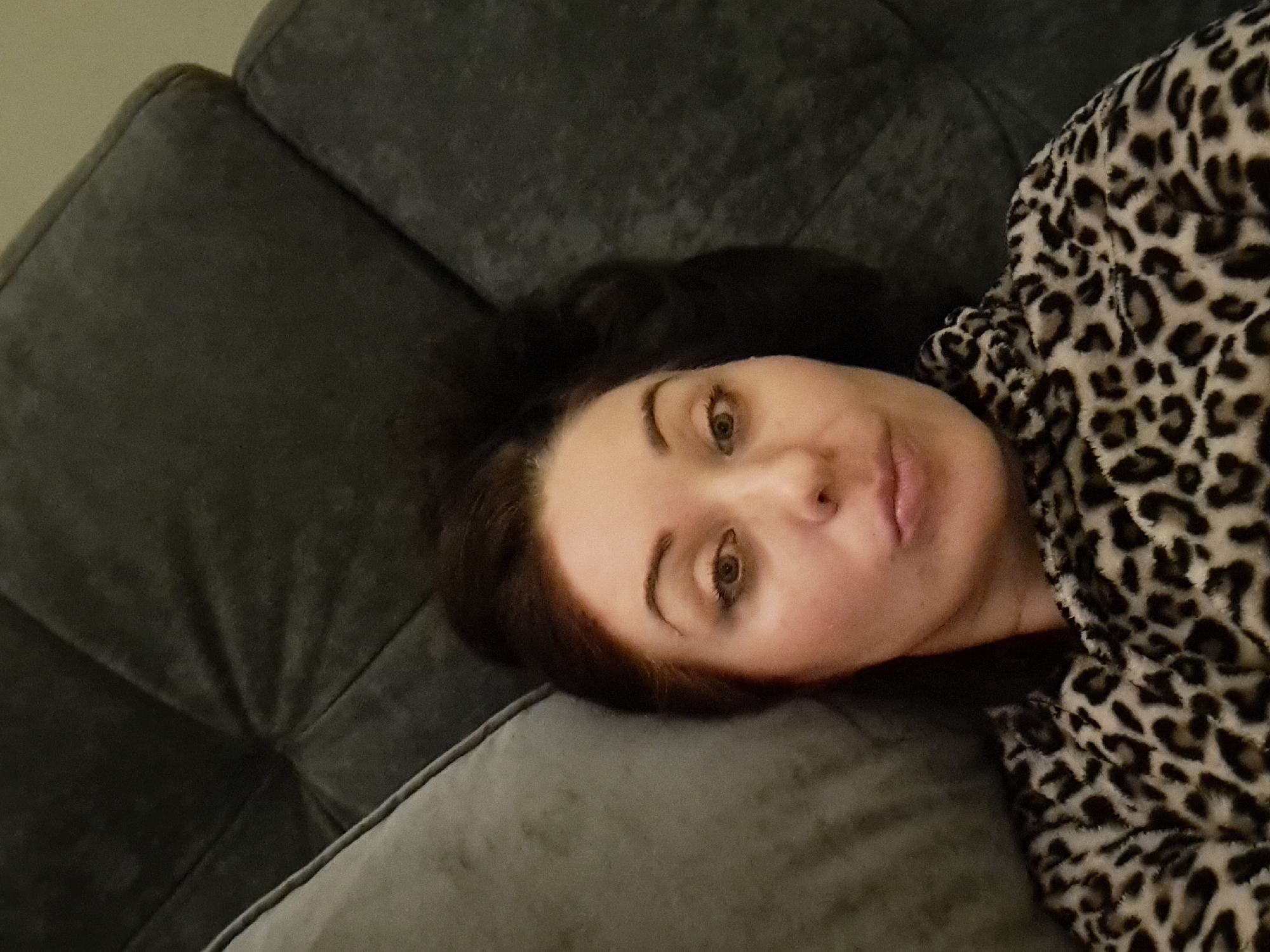 A woman is lying on a dark gray couch, with her head resting on a light gray pillow. She has dark brown hair, styled up. She is wearing a leopard print robe. The woman is looking directly at the camera with her lips slightly pursed. The background consists of the couch and a pillow. There is no indication of the location being Irkutsk, Russia.