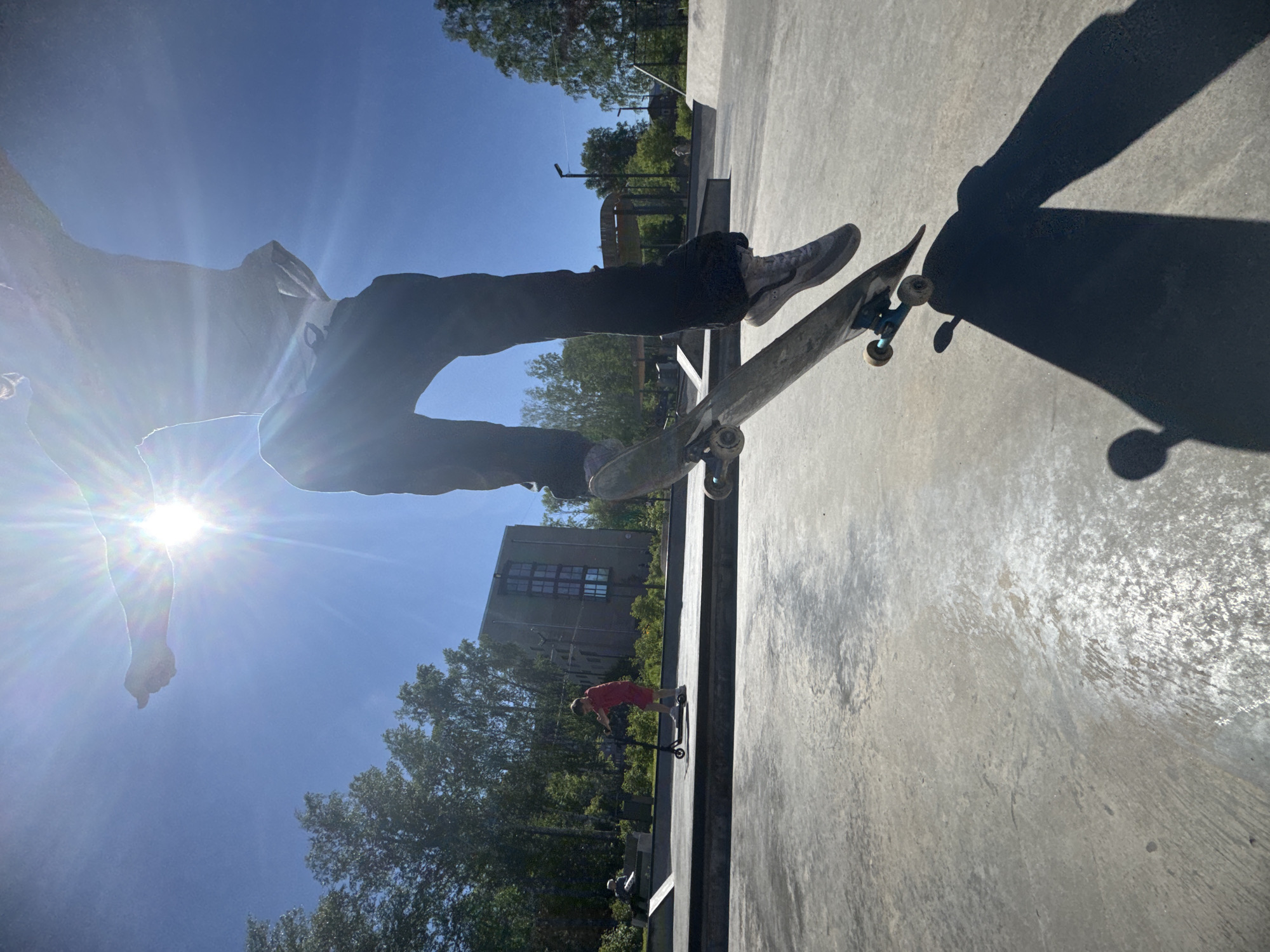 The image depicts a sunny day at a skatepark in Divnogorsk, Russia. The primary subject is a person mid-air on a skateboard, their body silhouetted against the bright sun. The skateboard is tilted, showing its wheels and trucks. In the background, a young boy in a red outfit rides a scooter. The skatepark is constructed of concrete with various ramps and structures visible. Trees and a modern building with a grid-like facade are situated behind the park, suggesting an urban or suburban setting. The sky is clear blue, and the sun's glare is prominent, casting a long shadow from the skateboarder onto the concrete ground.