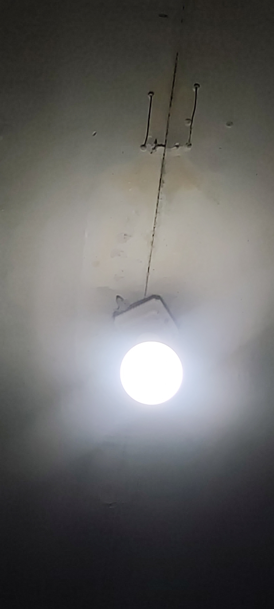 The image shows a bright, circular light source, likely an LED bulb, illuminating a ceiling. The ceiling appears to be made of concrete or plaster and has some visible imperfections and markings. A thin, dark line runs vertically down the center of the image, possibly indicating a joint or crack in the ceiling. Two sets of curved, dark lines, resembling staples or fasteners, are visible on either side of the central line. A white, rectangular object, possibly a light fixture or junction box, is partially obscured by the light's glare. The overall environment is dimly lit, with the bright light source dominating the center of the frame. The image does not provide any specific details or context that would allow for the determination of its location in the city of Karangwangun, Indonesia.