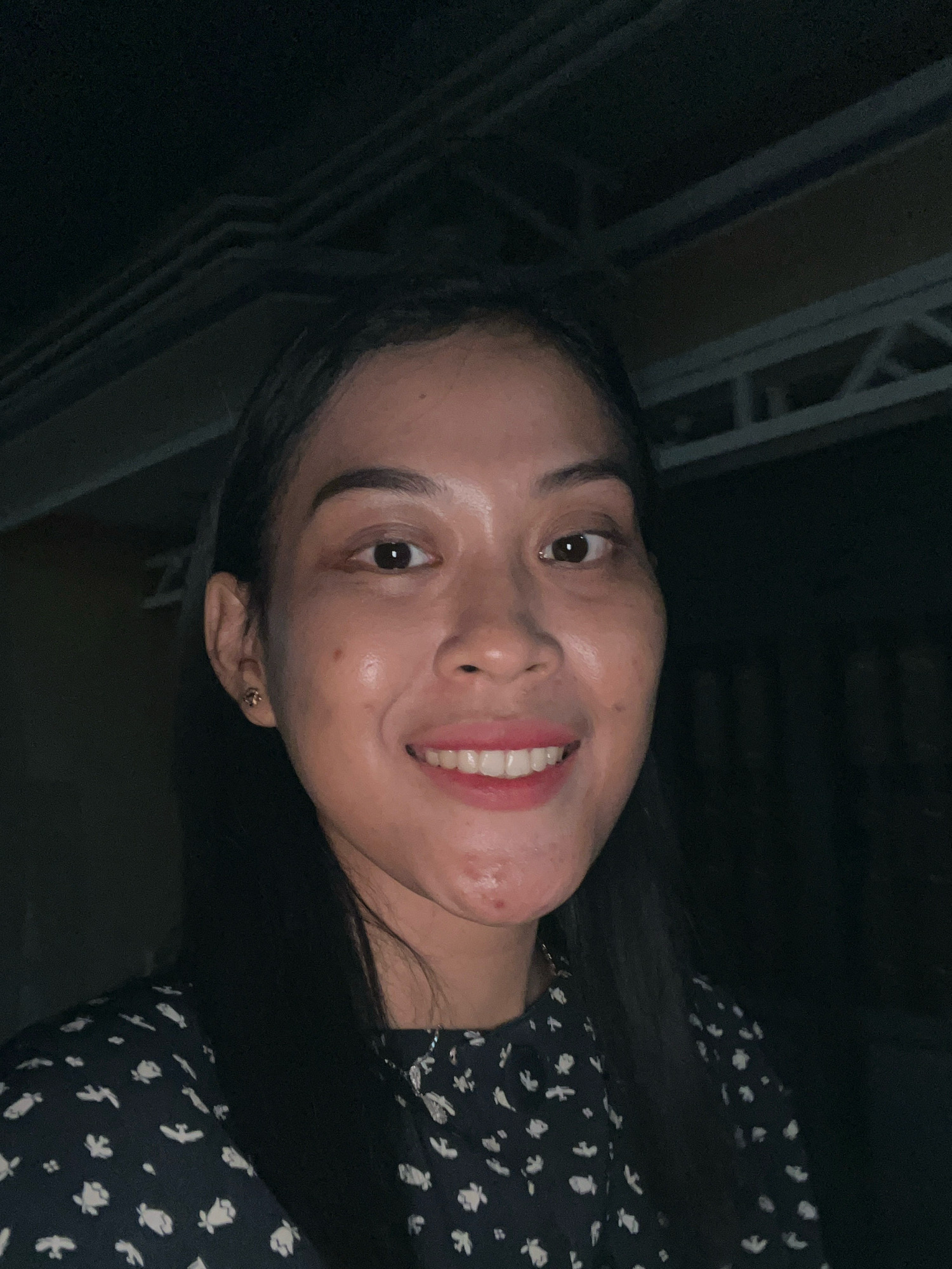 A close-up, low-light selfie shows a woman with dark hair and fair skin, smiling with her mouth slightly open, revealing white teeth and pink lipstick. She has a small gold earring in her left ear and visible blemishes on her chin. She is wearing a dark dress with a white floral pattern and a delicate silver necklace. The background is dark and out of focus, suggesting an indoor or evening setting. The overhead structure visible in the upper portion of the image appears to be a ceiling or roof with exposed beams, possibly in a building located in Kecamatan Pati, Indonesia.