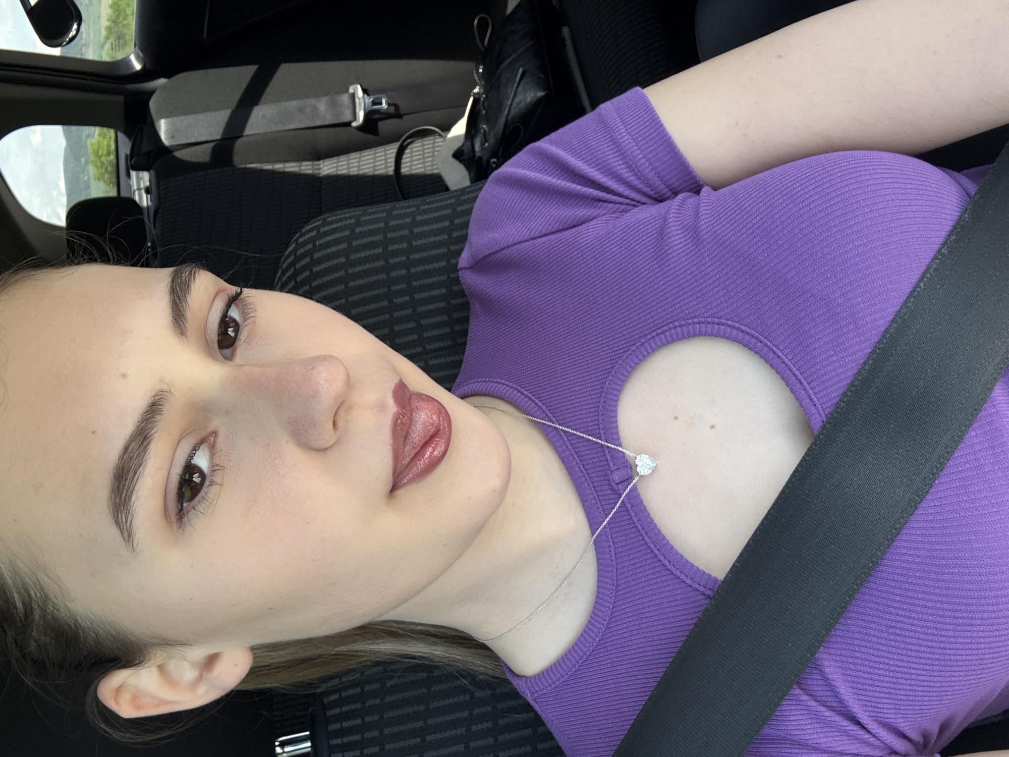 A fair-skinned young woman with brown eyes, light brown hair, and full lips is sitting in the passenger seat of a vehicle. She is wearing a purple ribbed top with a heart-shaped cutout at the chest. A silver necklace with a heart-shaped pendant is visible. A black seatbelt is fastened across her chest. The vehicle's interior includes dark grey patterned fabric on the seats. A glimpse of an exterior view, possibly trees or landscape, is visible through the vehicle's window. The location is not identifiable as the city of Levski, Bulgaria.