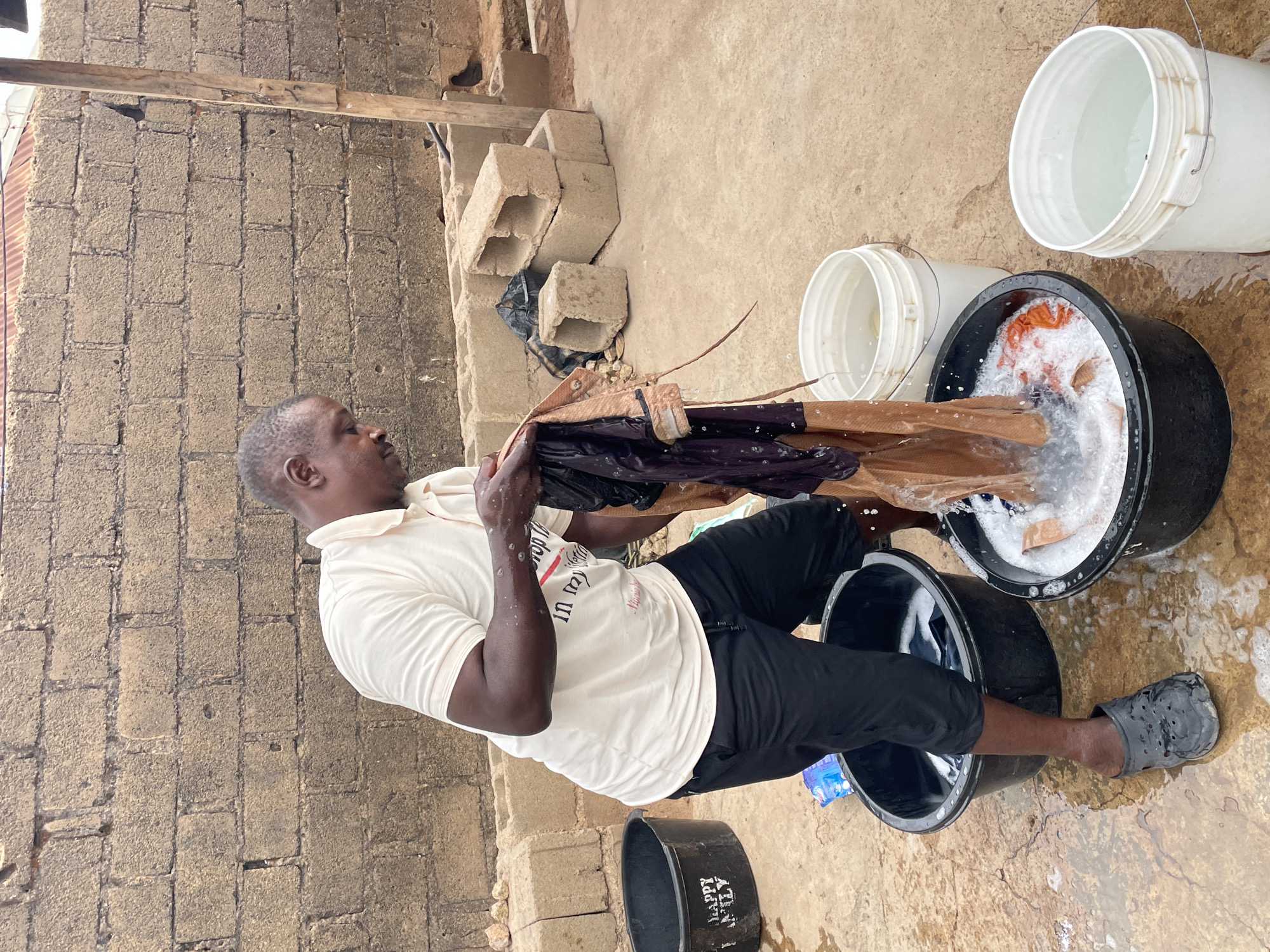 In Jos, Nigeria, a man is manually washing clothes outdoors. He stands between two black plastic tubs, one containing soapy water and clothes, the other seemingly empty. He is lifting a garment with both hands, seemingly inspecting it. The man wears a white short-sleeved t-shirt, black pants, and a single gray clog on his right foot. Three white plastic buckets are situated nearby, appearing to contain water. The washing area is outside, with a block wall in the background. Some concrete blocks are stacked near the wall.