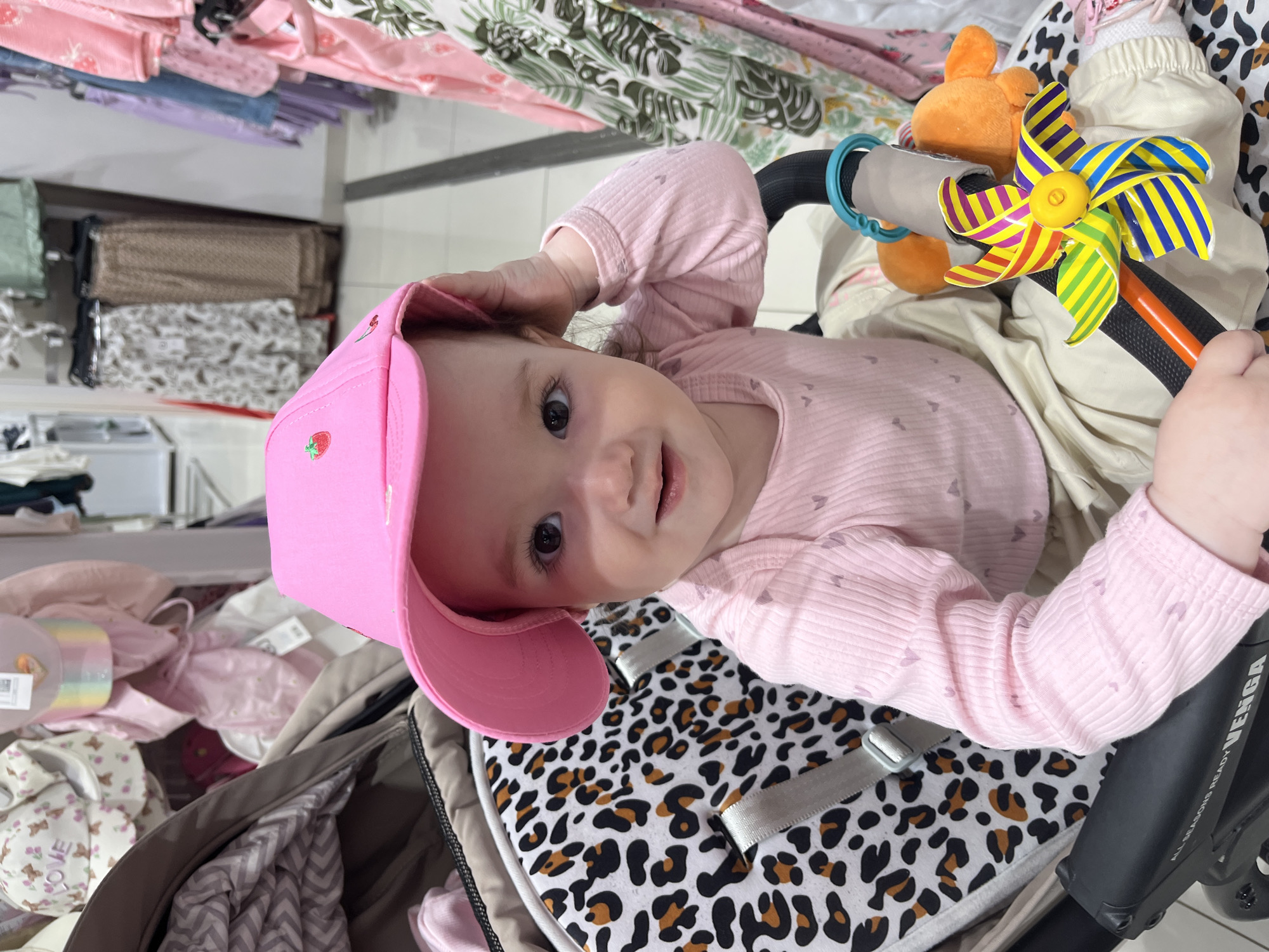 A baby wearing a pink cap with a strawberry embroidered on it is lying in a stroller with a leopard print seat. The baby is wearing a light pink long-sleeved shirt and is holding a colorful pinwheel toy. An orange stuffed animal is attached to the stroller's handlebar. Background elements include clothing racks with various garments, likely in a retail setting. The stroller has "VEGA" printed on its frame. The location is identified as Shushary, Russia.