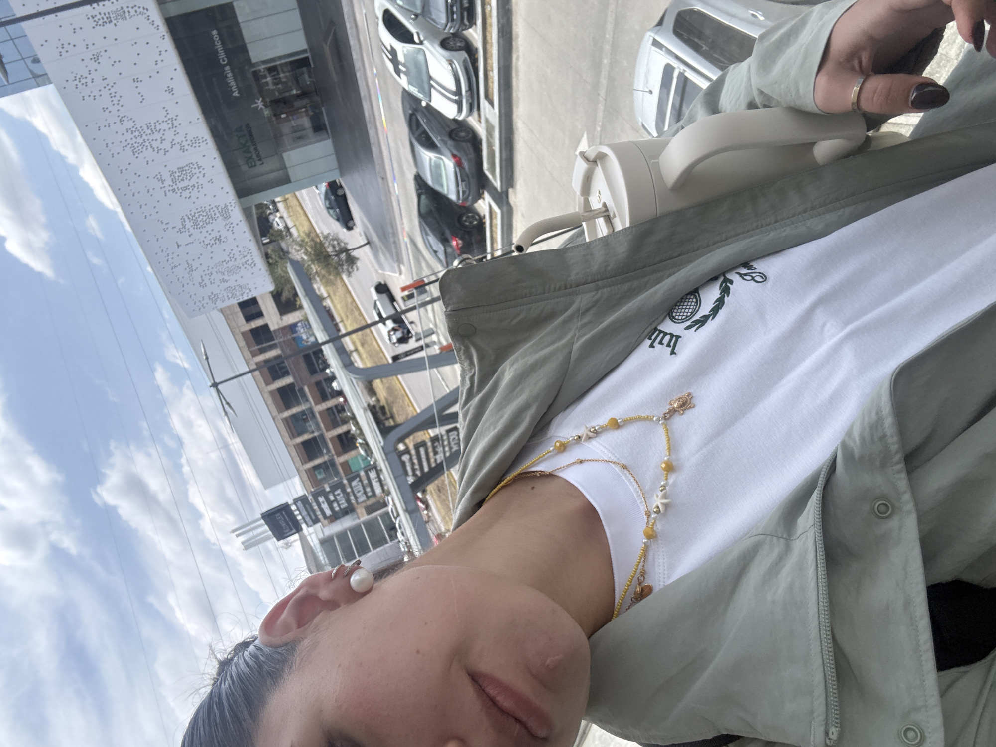 A medium-level shot shows a woman wearing a green jacket, white t-shirt, and jewelry, including a beaded necklace and pearl earrings. She has dark hair pulled back. Her face is visible, showing features like her eye, nose, and lips. In the background, there is an urban scene, including buildings, cars, and some greenery, indicating an outdoor setting. A white object, possibly a water bottle or container, is also visible. The image appears to be taken in the city of Puebla, Mexico, based on the visible signage "Análisis Clínicos".