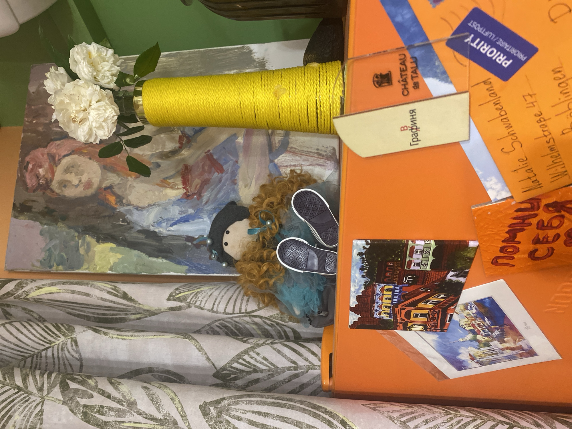 The image shows an indoor scene, possibly a room in Sochi, Russia, as indicated by the presence of a postcard featuring a building with architectural details common in the region. On an orange surface, likely a refrigerator, a doll with red curly hair, a black hat, and a teal dress sits next to a painting of a woman with a red headscarf. Beside the painting, a yellow yarn-wrapped vase holds white flowers. To the left, a curtain with a leaf pattern is visible. Several items are attached to the orange surface: a small canvas painting of a European-style building, a framed print of a European cityscape, a torn piece of paper with Russian text that reads "ПОМНИ О СЕБЯ" (Remember Yourself), and two other paper items, one with Russian text and a logo for "CHÂTEAU de TALU," and another with handwritten address details for "Natalie Schwabenland Wilhelmstraße 47 Böblingen" and a blue "PRIORITY PRIORITAIRE/LUFTPOST" sticker. The overall impression is one of a decorated, personal space.