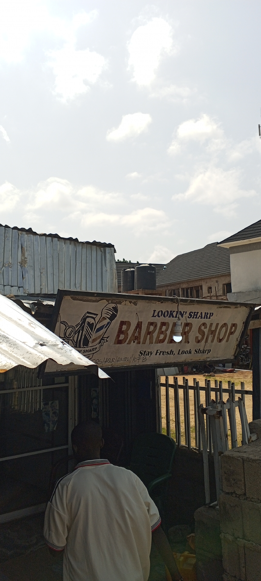 The image depicts a street scene in Jalingo, Nigeria, featuring a barber shop with a prominent sign. The sign, partially weathered, reads "LOOKIN' SHARP BARBER SHOP" with the tagline "Stay Fresh, Look Sharp". A graphic of a barber's clippers and scissors is visible on the left side of the sign. Beneath the main text, there appears to be an inscription of letters and numbers, possibly a contact or address detail, including "HQR/HMRH/107B".
In the foreground, a man is seen from the back, wearing a light-colored short-sleeved shirt with red and blue striped trim on the collar and sleeves. He is standing near the entrance of the barber shop. The shop itself appears to be a simple structure with a corrugated metal roof and possibly some fencing.
In the background, several dark-colored water tanks are visible on top of a building. Further back, the roofs of other buildings, constructed with dark tiles and concrete, suggest an urban or semi-urban environment. The sky is overcast with bright white clouds. The overall impression is of a functional, local business operating in a typical Nigerian town setting.