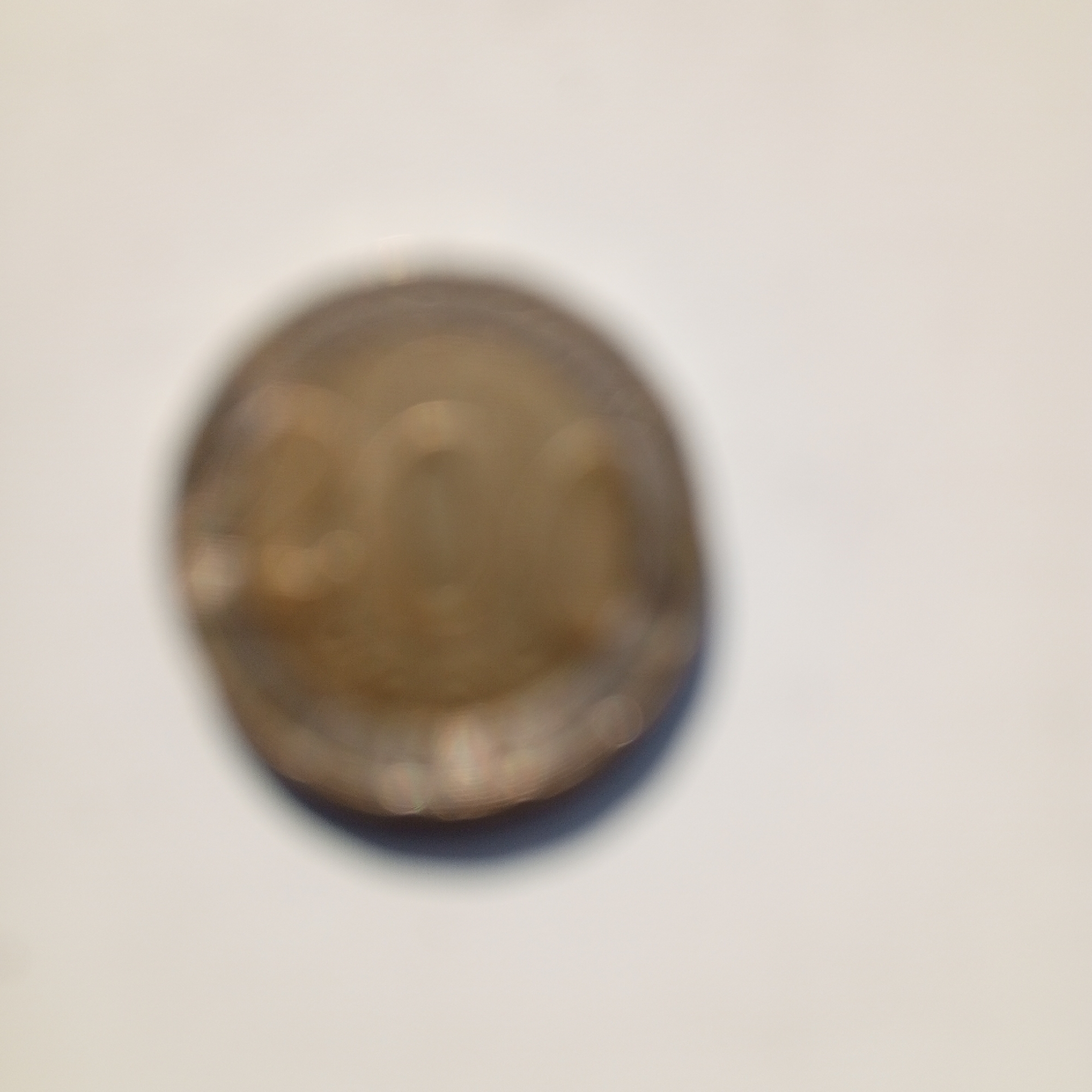 The image shows a blurred close-up of a coin against a plain, light-colored background, likely a table or surface. The coin is round and metallic, with visible concentric circular patterns suggesting it is a minted coin. The colors are muted, with shades of gold, brown, and hints of grey. Due to the blurriness, specific details like markings, text, or the denomination of the coin are not discernible. The lighting creates some bright spots on the coin's surface, indicating a light source from the side or above. There is no indication of the coin's location in Sayanogorsk, Russia, or any other specific geographical context.