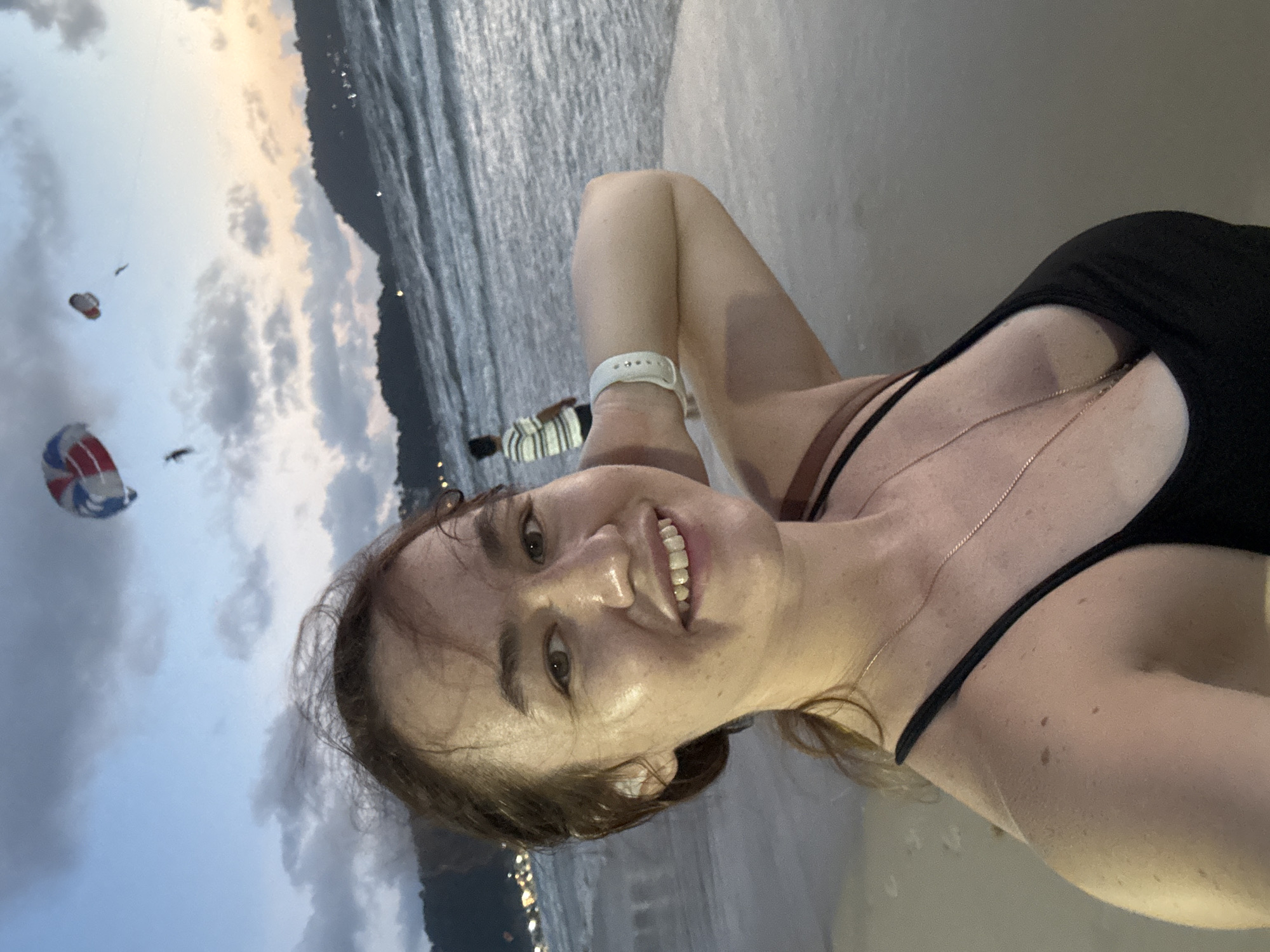 A woman with brown hair tied in a ponytail, wearing a black tank top and a necklace, is taking a selfie on a beach in Patong, Thailand. She is smiling at the camera, with her right arm bent at the elbow and her hand behind her head. The ocean is behind her, with waves breaking on the shore. In the sky, there are two paragliders. In the background, there is a coastline with hills and lights from the town. The sky is a dusky blue with clouds and hints of orange and yellow from the setting sun.