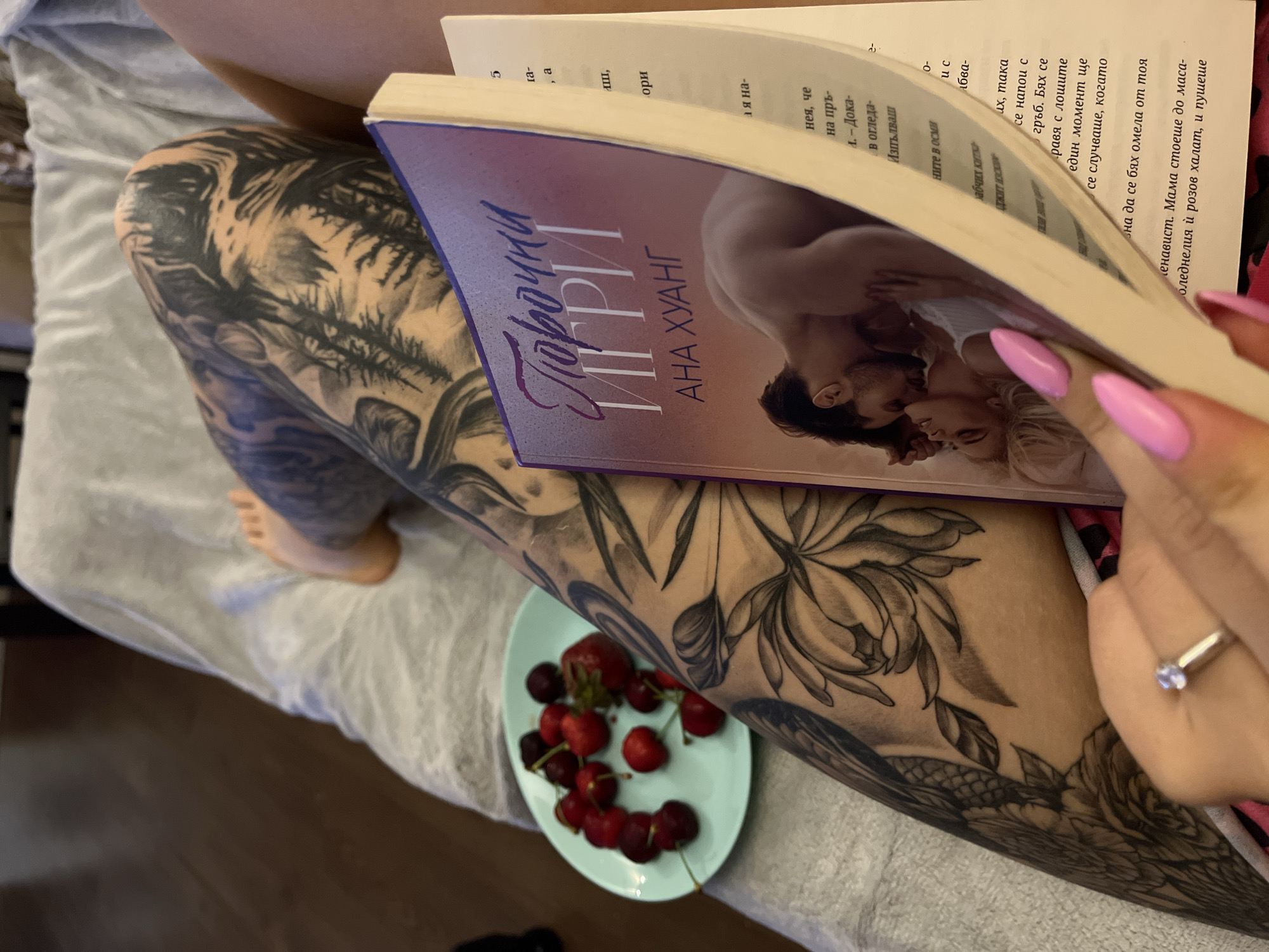 An overhead, close-up shot captures a person relaxing indoors, likely in Veliko Tarnovo, Bulgaria, engrossed in reading. The person's legs, extensively covered in intricate black and grey tattoos depicting forest scenes and large floral designs, are central to the frame. One leg rests on light gray, textured bedding or a blanket, while the other is slightly bent.
Hands with long, bright pink, almond-shaped nails hold an open book. A silver ring with a single light-colored stone adorns the ring finger of the left hand. The book's partially visible purple cover features a romantic illustration of an embracing couple, with the Bulgarian title "Порочни ИГРИ" (Vicious Games) and author "АНА ХУАНГ" (Ana Huang) clearly displayed in Cyrillic. The open pages also show Cyrillic text, with snippets like "Мама стоеше до маса-" and "розов халат" visible, confirming the language.
Positioned on the bedding near the tattooed leg is a small, light blue round plate holding a fresh snack of several red cherries with stems and one strawberry. The scene suggests a moment of leisure and relaxation. The floor visible beneath the bedding is dark.