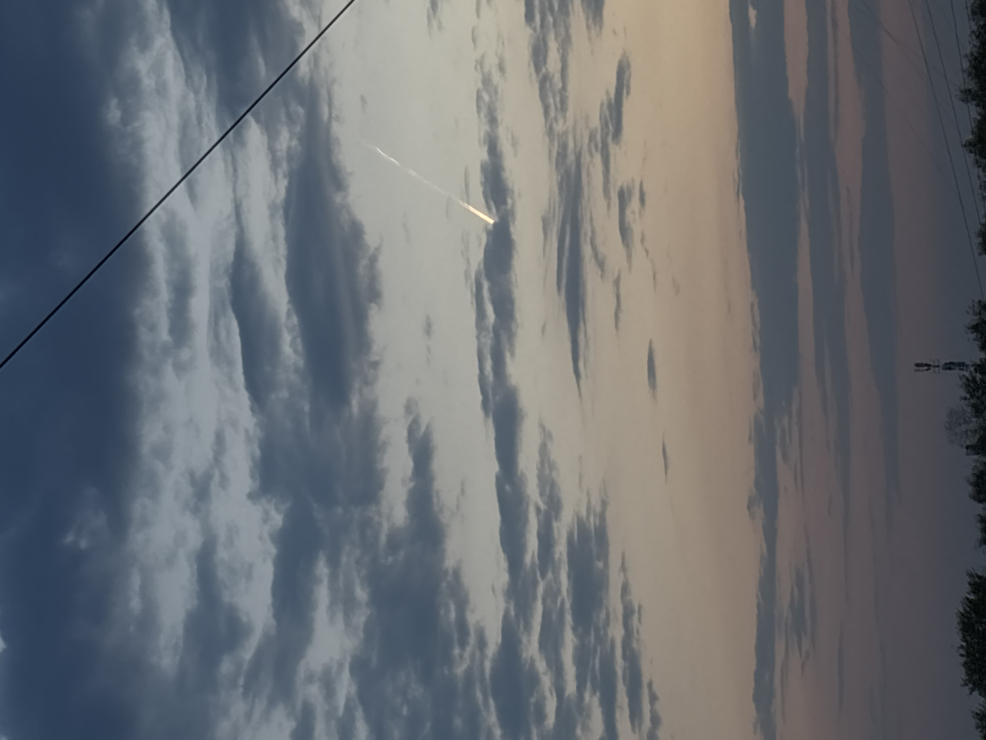 The image captures a sky with varied cloud formations, ranging from dark, dense clouds to lighter, more diffused ones. A distinct contrail is visible, indicating the passage of an aircraft. An overhead cable cuts across the upper left corner. On the far right, there are trees, powerlines and power towers visible. The location is Chaikovsky, Russia.