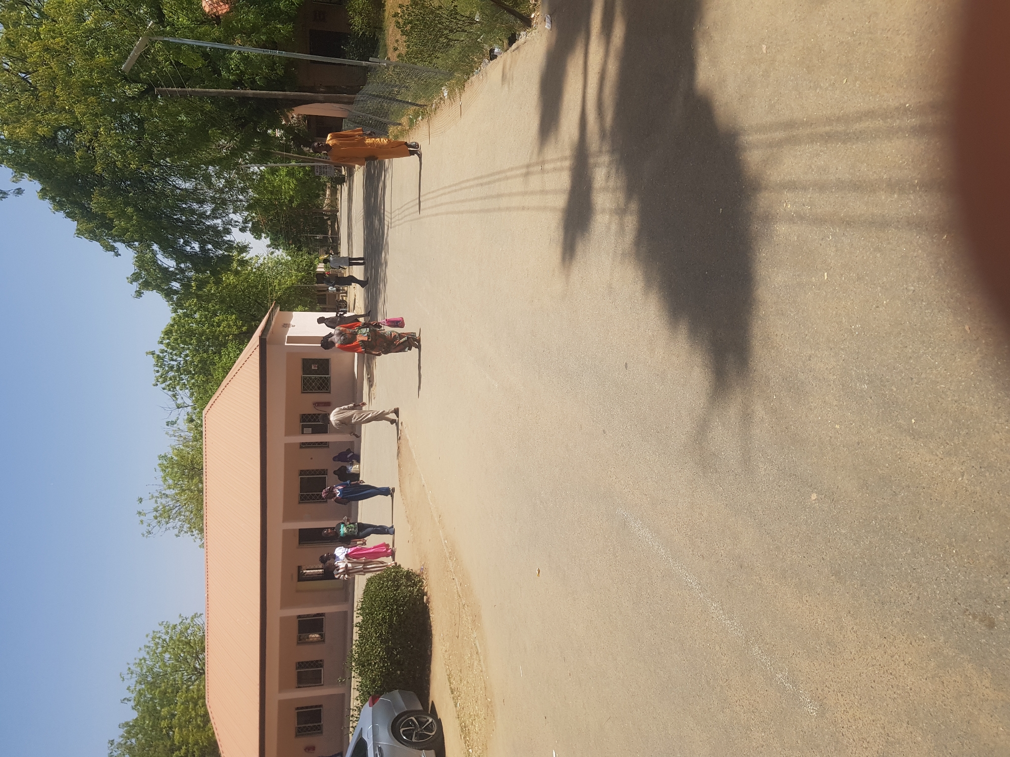 The scene is a street in Dabawa, Nigeria. A one-story building is on the left, with a peach-colored roof and walls. People are walking along the side of the building, some standing near the entrance. A gray car is parked near a bush to the right of the building. Overhead power lines run along the street, casting shadows on the pavement. A person in a brown robe is walking away in the distance. There are trees on either side of the road. The sky is blue.