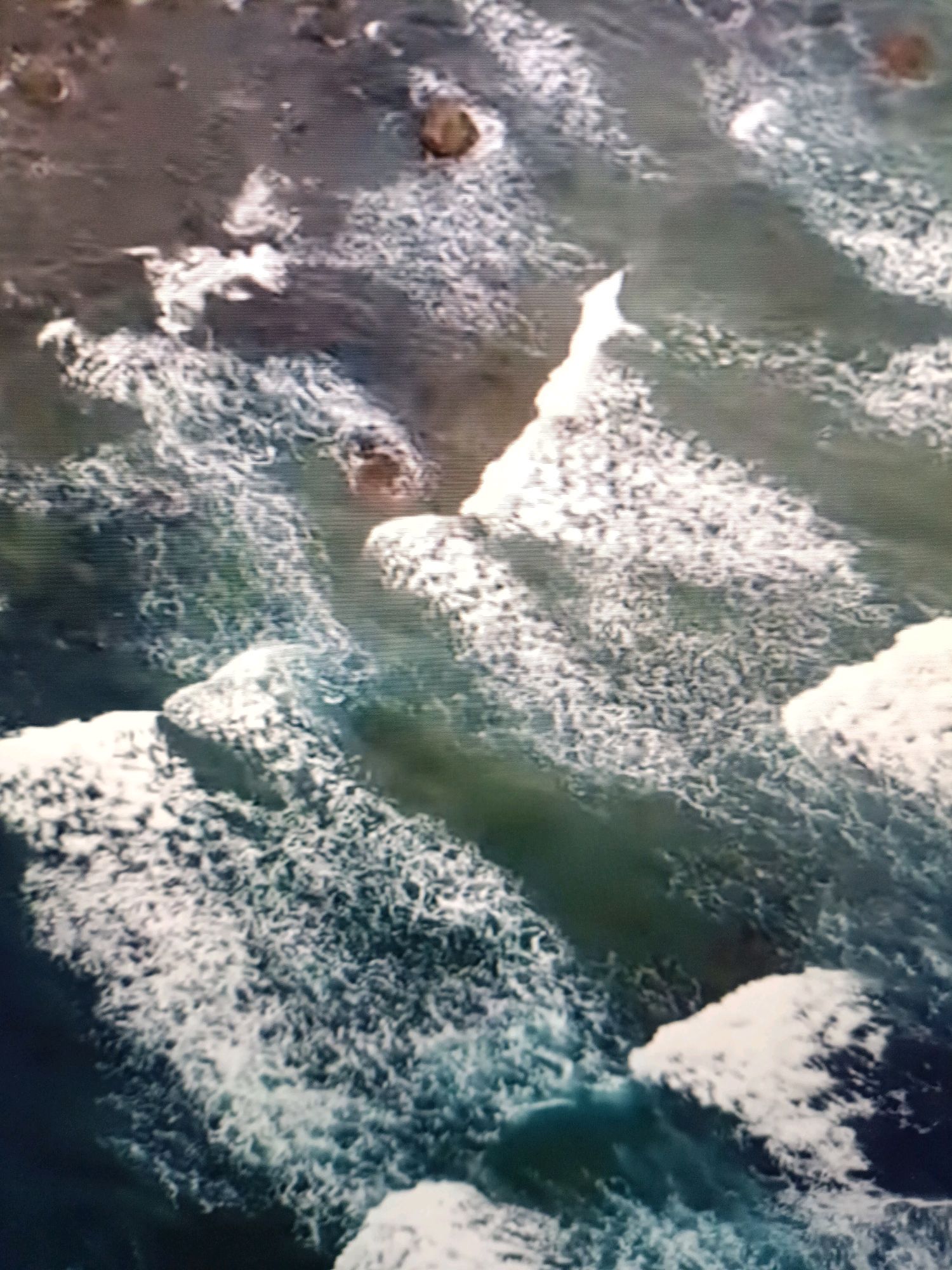 This image is an aerial view of waves crashing against rocks along the coast of Netanya, Israel. The water is a deep blue-green, and the waves are white and foamy as they break. There are several large rocks visible in the water, some of which are partially submerged. The shoreline appears to be rocky and uneven. The image is somewhat blurry and pixelated, suggesting it may be a low-resolution photograph or video still. The scene depicts a natural coastal environment with no signs of human activity or artificial structures.