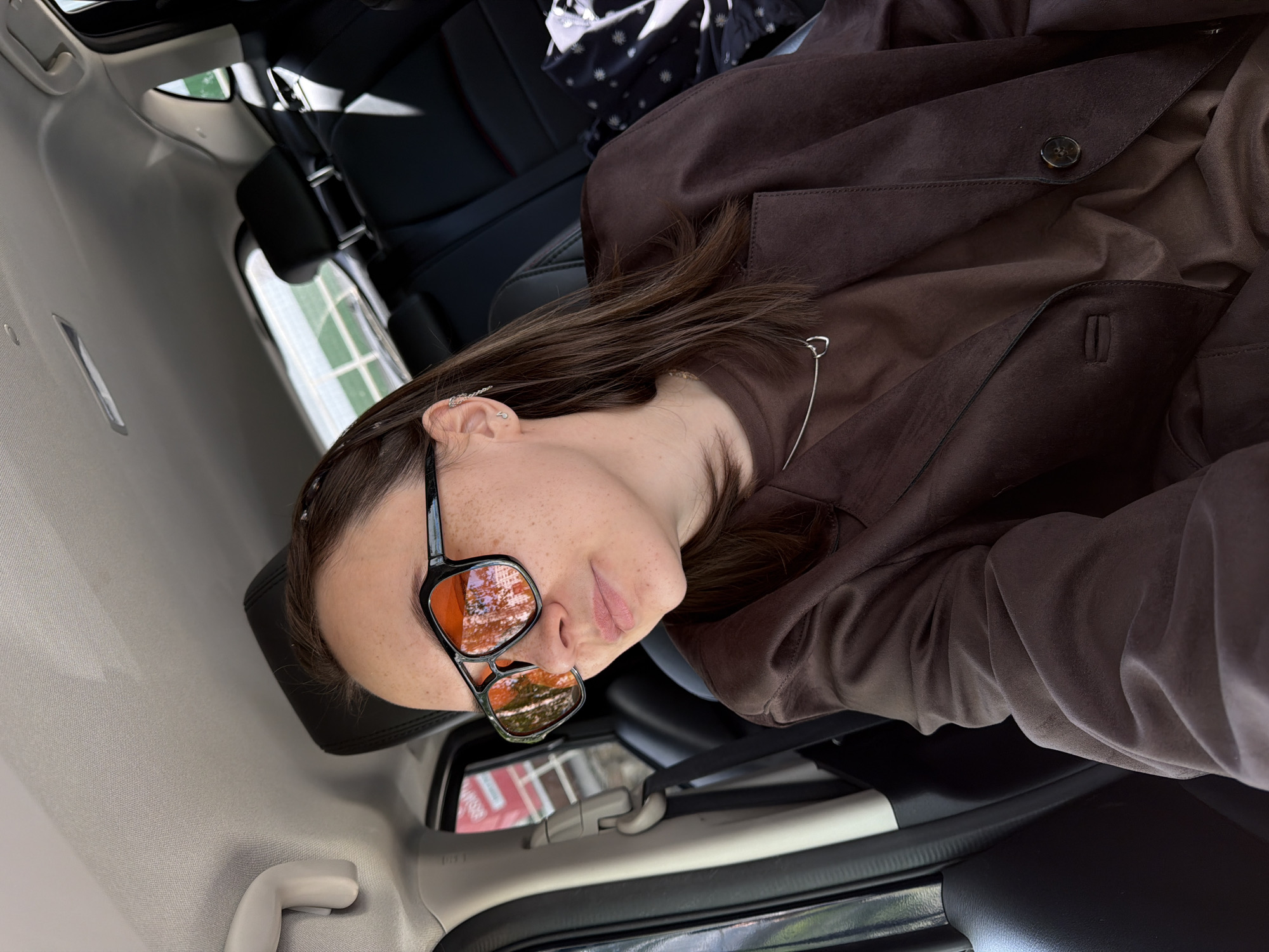 A young person with freckles is lying inside a car, wearing sunglasses that reflect the exterior environment. The person is wearing a brown top and jacket. The car's interior features black leather seats, and the view through the windows suggests the location is likely in a residential area in the city of Chaikovsky, Russia, based on the reflection in the sunglasses, showing a red and white building across the street. The photograph is taken from an unusual angle, possibly from the side of the subject.