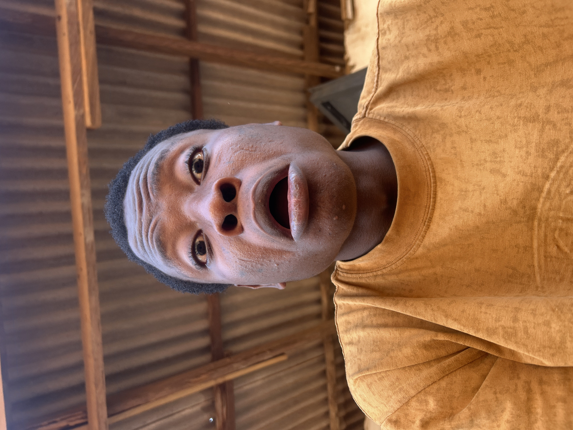 A close-up, low-angle shot captures a person with their mouth open in apparent surprise or shock. The person has dark hair and is wearing a golden-yellow t-shirt. The environment includes corrugated metal and wooden beams, suggesting the location might be a simple, possibly rustic, structure in Unknown, Unknown.