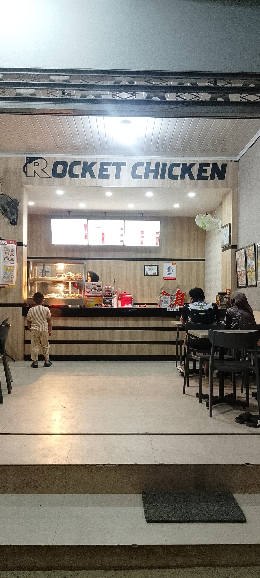 The image depicts the interior of a fast-food restaurant named "ROCKET CHICKEN" in Batulicin, Indonesia. The restaurant has a clean and modern aesthetic with wood-paneled walls and a white slatted ceiling. The main counter area features a glass display case showcasing fried chicken, a cash register, and various condiments. Three digital menu boards are mounted above the counter, displaying prices and offerings in red text on a white background.

In the foreground, a young boy wearing a striped jumpsuit stands facing the counter, appearing to be a customer. To the right, two individuals are seated at tables. One person, wearing a black shirt with white lettering, faces away from the camera, while the other, wearing a hijab, is also seated at a table.

The environment is brightly lit by overhead fluorescent lights. A ceiling fan is visible on the right wall, and a wall-mounted fan is present on the left. The floor is tiled with a light-colored ceramic material. In the lower portion of the image, a dark grey mat is placed on a step leading down from the restaurant's entrance area. Various promotional posters and signs are visible on the walls and near the counter. The overall scene suggests a typical operational environment for a fast-food establishment.