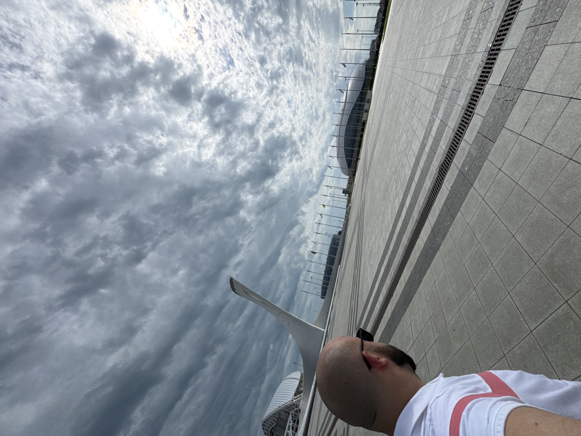 The photograph shows an outdoor scene in the city of Sirius, Russia. The sky is overcast with heavy clouds. In the middle ground, the Fisht Olympic Stadium and a tall, white, curved structure are visible. Rows of flagpoles line the area between the stadium and a wide expanse of paved tiles. A drainage grate runs along one edge of the tiles. In the foreground, the back of a person's head is visible. The person has dark hair and is wearing a white shirt with pink suspenders. The photograph is taken at an angle, tilting the horizon.