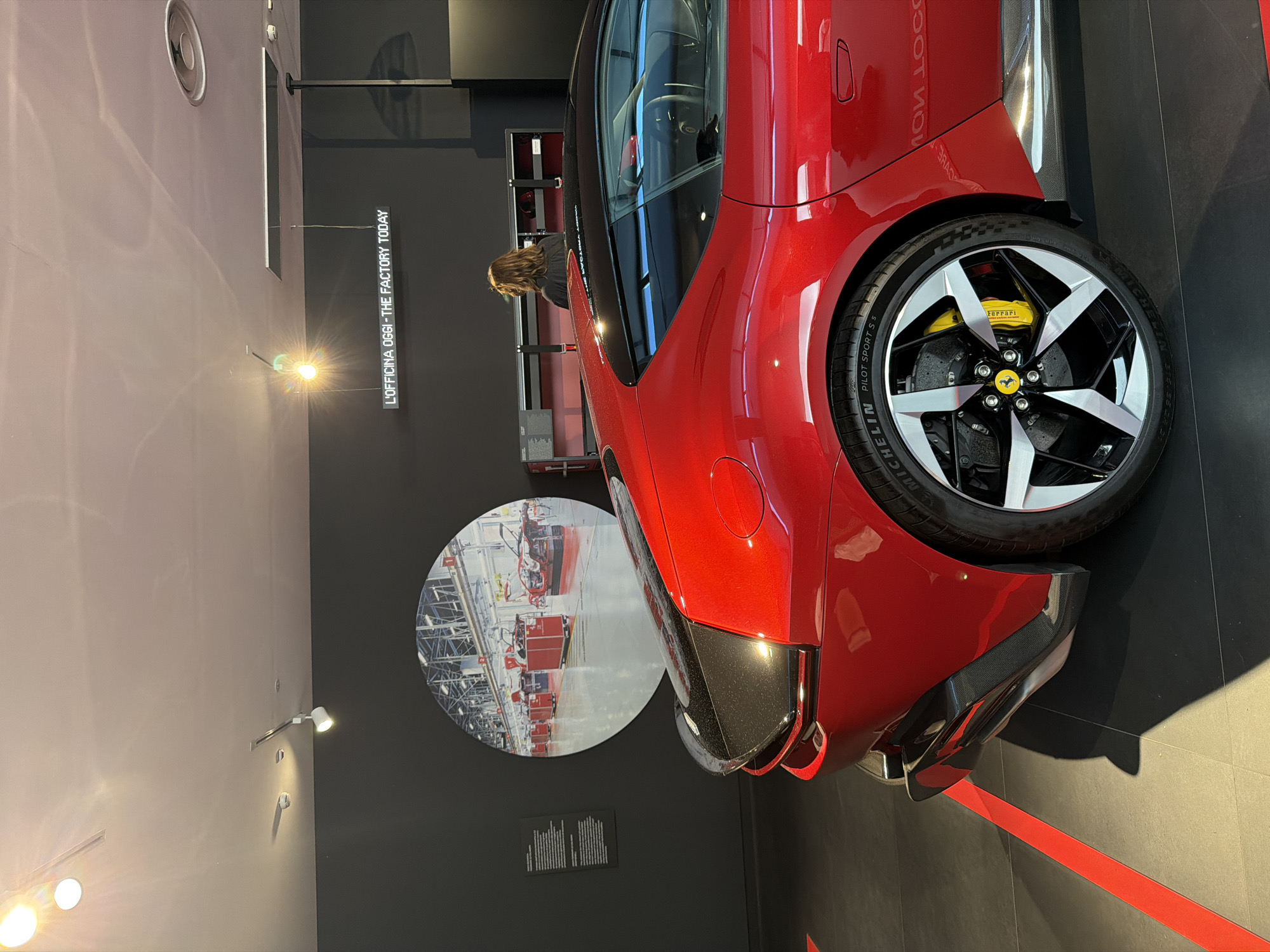 The image depicts the interior of a modern showroom, likely located in Maranello, Italy, given the prominent Ferrari branding. A bright red Ferrari car, viewed from the rear quarter panel, is the central subject. The car features a black carbon fiber rear spoiler and a distinctive wheel with silver and black spokes, yellow brake calipers, and a Michelin Pilot Sport tire.
Behind the car, the wall is dark gray. To the left, a large circular graphic displays an image of a factory floor with red trolleys. To the right of the graphic, a digital display on the wall reads "L'OFFICINA OGGI - THE FACTORY TODAY." A person, whose back is to the camera, is standing and looking at a display case filled with what appear to be automotive parts or memorabilia. The floor is dark, with bright red lines forming geometric patterns. Track lighting illuminates the space from the ceiling, casting shadows. The overall environment suggests a high-end automotive exhibition or a brand experience center.