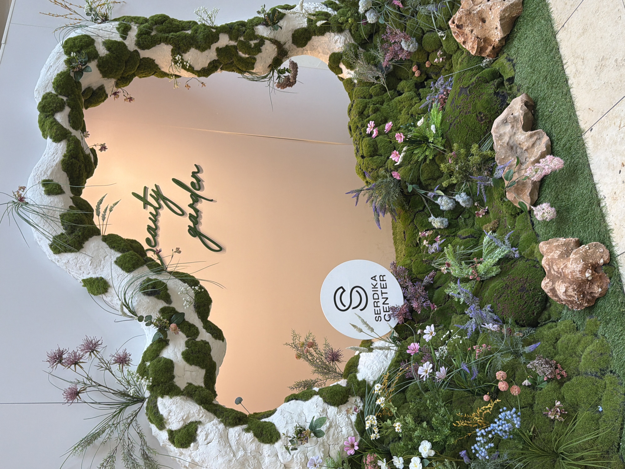 The image depicts a decorative installation that includes a large, irregular, heart-shaped frame constructed of a white, textured material with green moss patches, positioned against a light-colored wall. Inside the frame, the words "beauty garden" are displayed in dark green lettering. A white circular sign with black stylized "S" logo and text "SERDIKA CENTER" is positioned at the bottom right of the frame. The surrounding area is decorated with various types of green moss, flowers in shades of white, pink, purple, and blue, and some brown rocks. The top right corner also features an area covered in artificial green grass. The image likely depicts an installation at the Serdika Center shopping mall in Sofia, Bulgaria.