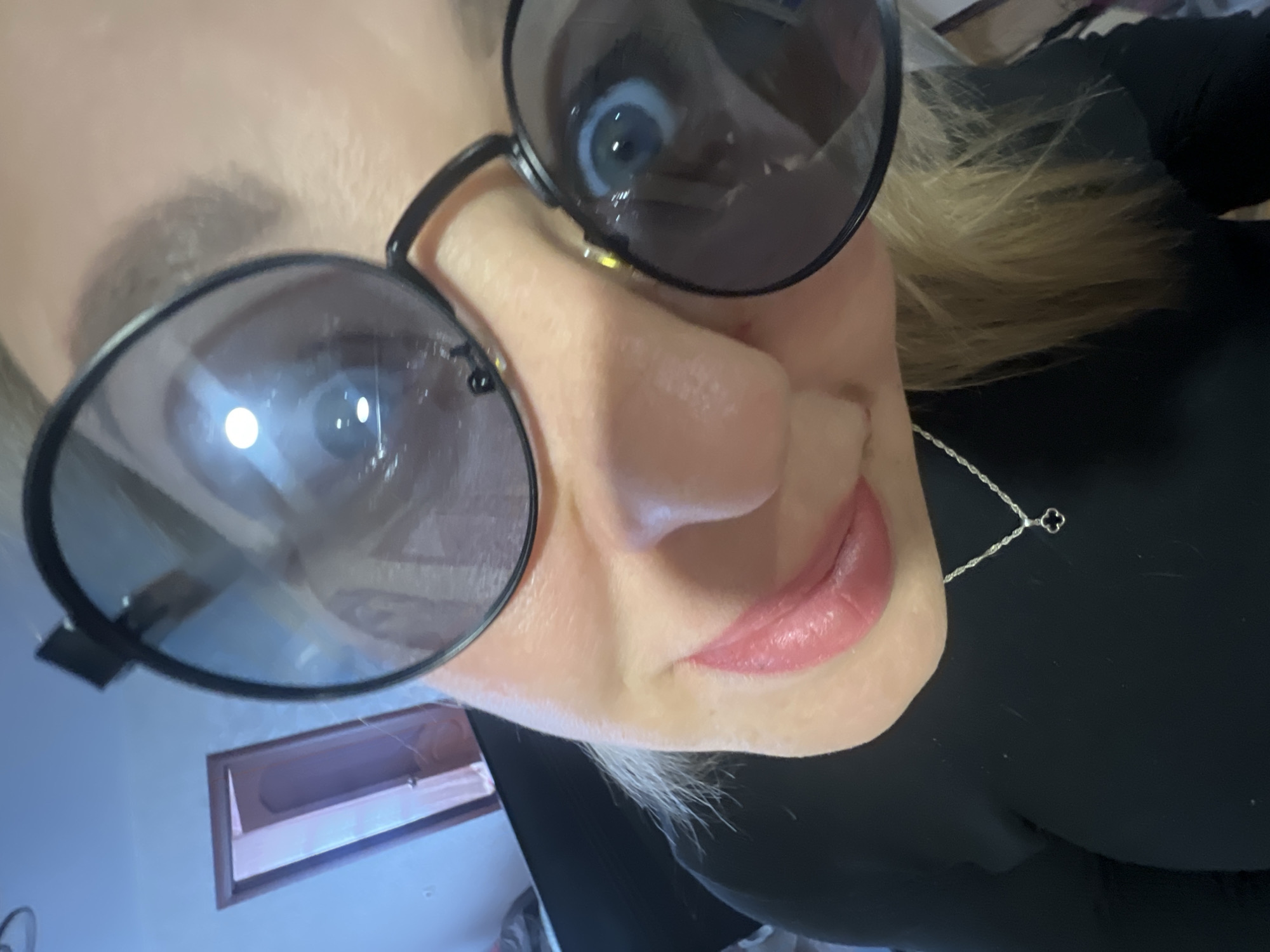 A selfie was taken at an angle, close-up of a fair-skinned individual with light blonde hair, wearing a black top and oversized sunglasses with a black frame. A silver necklace with a small pendant is visible around their neck. The reflection in the sunglasses suggests an indoor environment. In the lower left corner of the frame, a window or picture frame is visible on a light blue wall. The setting does not provide enough information to indicate if it is the city of Chaikovsky, Russia.