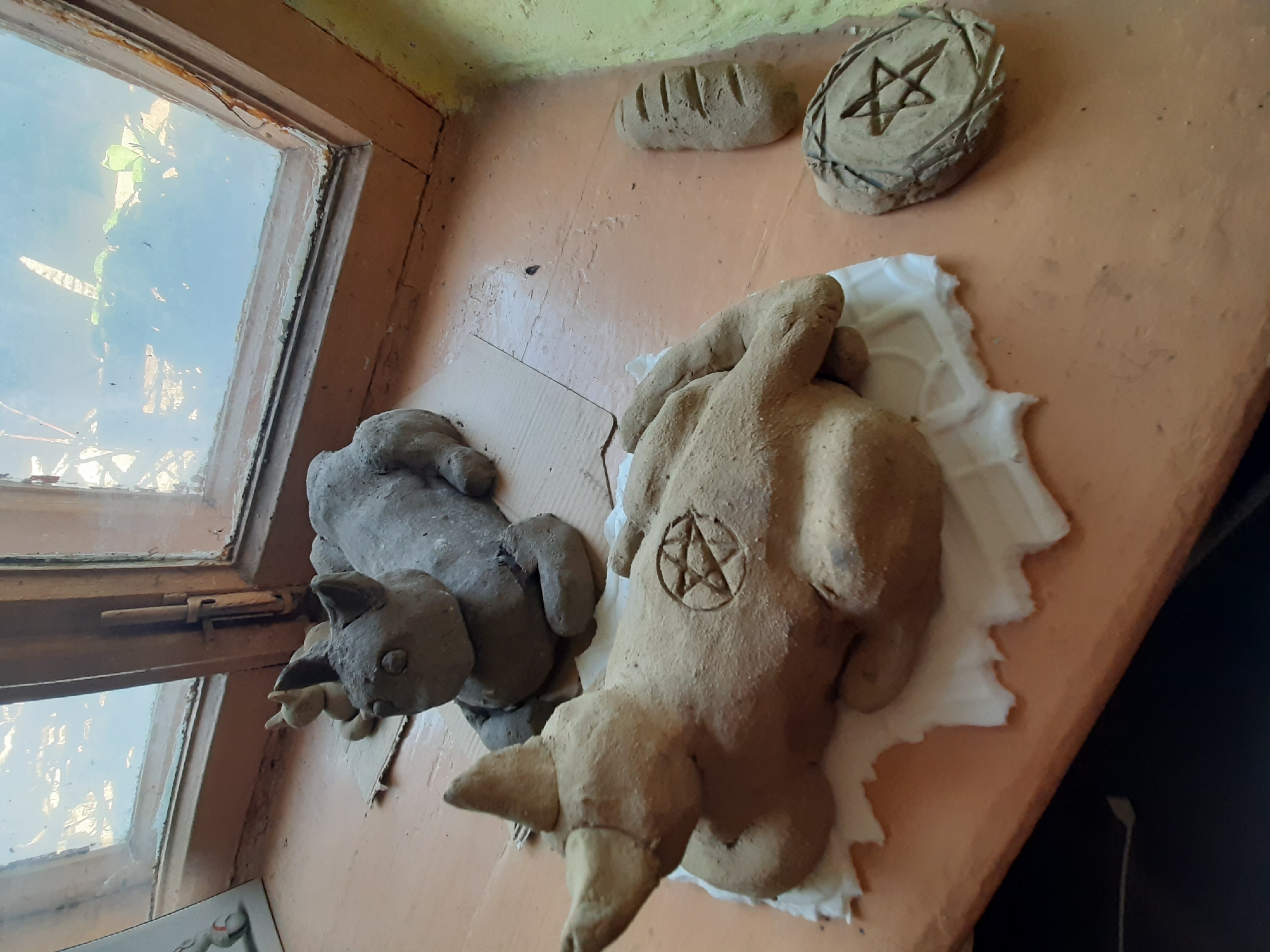 The image shows two clay sculptures on a wooden surface, possibly a windowsill or shelf. To the left, a gray, cat-like figure with large ears and a smaller kitten figure are placed near a window with a damaged wooden frame. The windowpane is reflective, showing a blurry outdoor scene with green foliage. To the right, a tan-colored sculpture, possibly representing a goat or devil, lies on a white piece of fabric. This sculpture has prominent horns and a pentagram symbol etched into its torso. Further to the right, two smaller clay objects are visible: one is oblong with incised lines resembling a loaf of bread, and the other is oval with a five-pointed star within a circular border, also incised. The wooden surface appears worn and has an orange-brown hue. The wall above is painted a pale yellow. The scene is indoors, with natural light coming from the window, suggesting it is daytime. The overall impression is one of an artistic or perhaps ritualistic arrangement. No visible text is present in the image. The location is identified as Bilhorod-Dnistrovskyi, Ukraine.