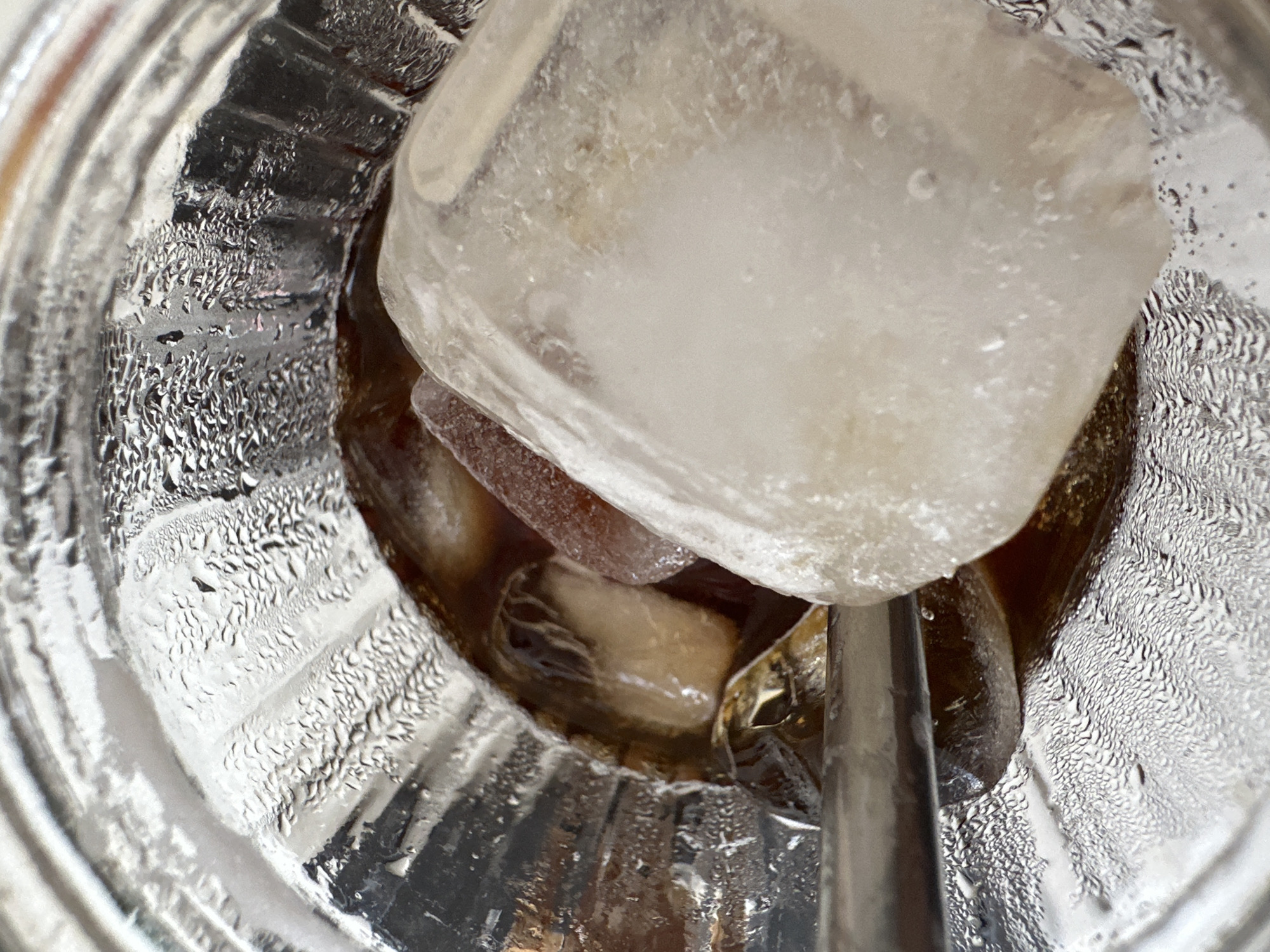 A close-up, overhead view shows a glass filled with ice and a dark liquid, likely coffee or iced tea. A large ice cube dominates the upper portion of the frame, partially submerged. Several smaller ice cubes and pieces are visible in the liquid below, with some appearing to be melting. The exterior of the glass is covered in condensation, indicating a cold beverage on a potentially warm day. The glass appears to be made of clear, ribbed material. A silver straw or stirrer is partially visible in the lower right quadrant of the image, submerged in the drink. The lighting suggests it is daytime, though no external environmental cues are present to determine the exact time of day. The scene is set indoors, in a casual setting, possibly a home or cafe. No people are visible, and no text is present in the image. The overall impression is one of a refreshing, cold drink.