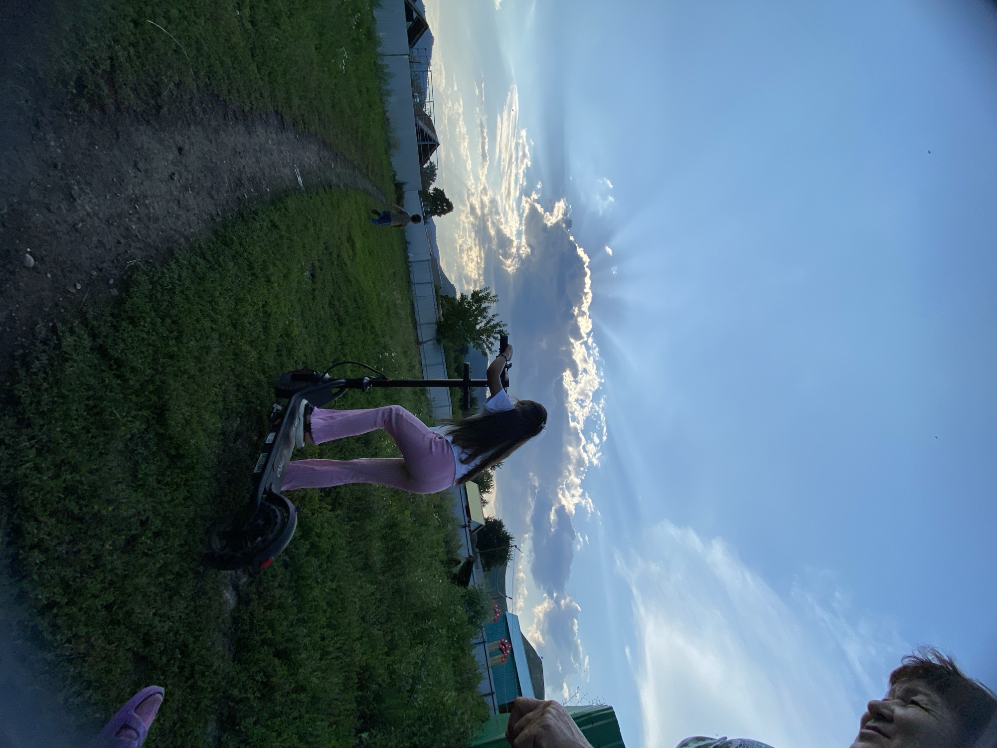 An outdoor scene in Maloarslangulovo, Russia, captures a cloudy sky with crepuscular rays emanating from behind a large cloud. In the foreground, a person is riding an electric scooter. To the left, a dirt path winds through a grassy field. Further in the background, a young boy is running along the path. A white fence runs along the edge of the property, behind which are several houses and trees.