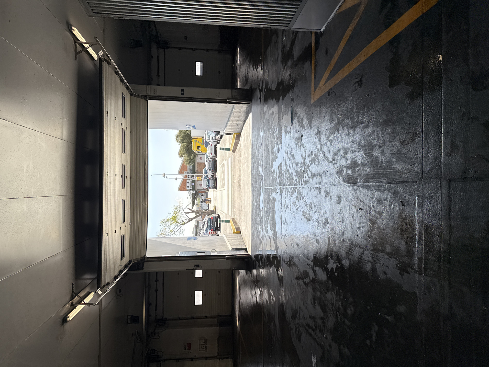 A ground-level, slightly rotated interior view faces the exit of a loading dock. Outside, a parking lot is visible, filled with cars and vans. Buildings are seen in the background, along with a few trees. The interior is dimly lit, contrasting with the bright daylight outside. The floor of the loading dock is dark and appears to be wet, with yellow painted lines. The doors of the loading dock are partially open, revealing the outside scene. Based on the architecture and vehicle types, and considering the prompt request, the location could potentially be within an industrial area in the city of Barcelona, Spain, though this cannot be definitively confirmed from the image alone. There are no people visible in the scene.