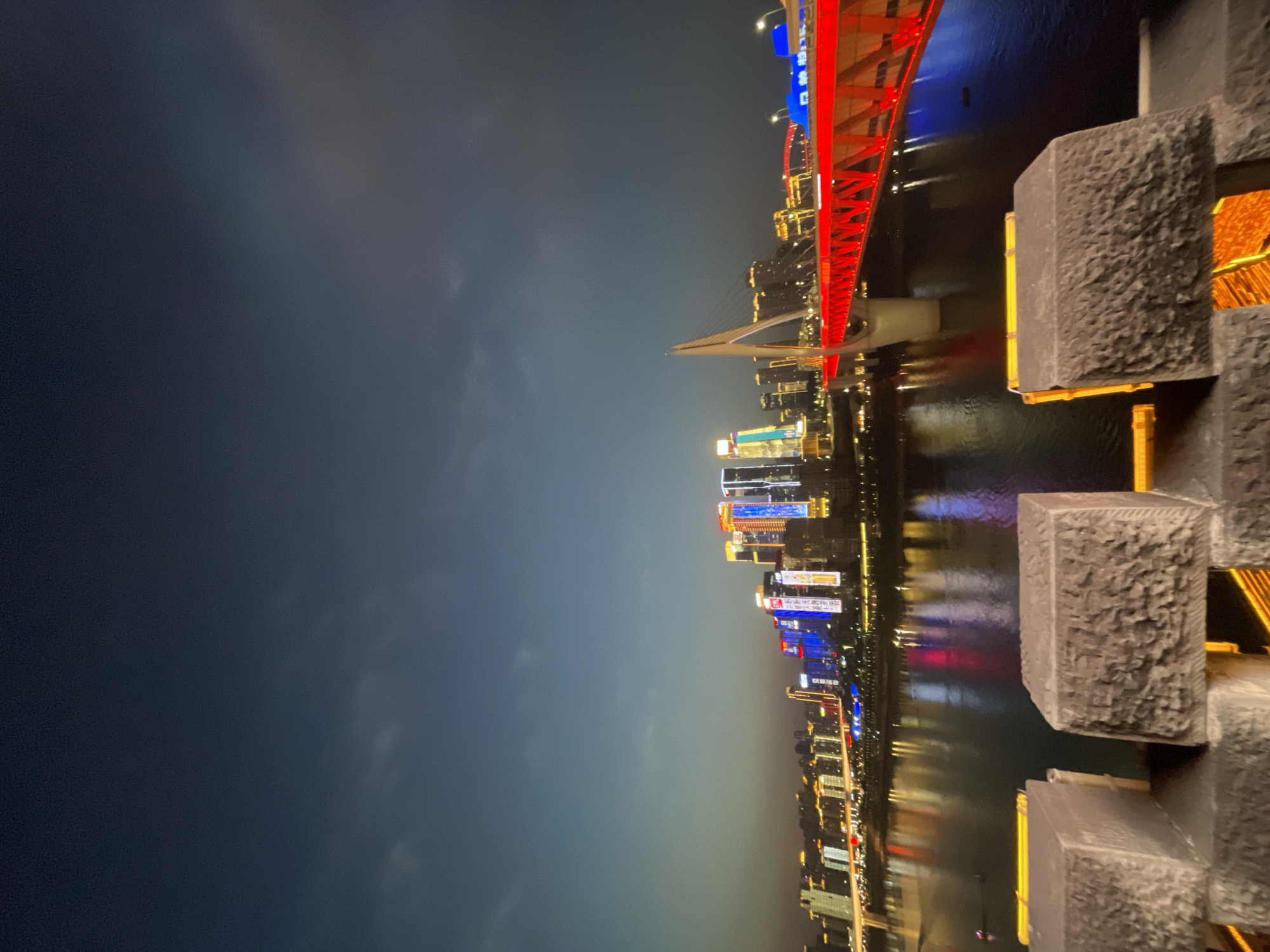 The image captures a night view in the Yuzhong District of Chongqing, China. A red steel arch bridge with blue and white lighting spans a river. Reflections of the bridge and city lights shimmer on the water's surface. In the foreground, there is a decorative structure constructed of stacked gray stone blocks with internal lighting casting a warm glow. The background is dominated by a dark sky. The city skyline is visible on the right side of the frame, featuring a dense collection of illuminated skyscrapers with a variety of colors. The image appears to be taken from an elevated perspective. No people are visible in the scene.