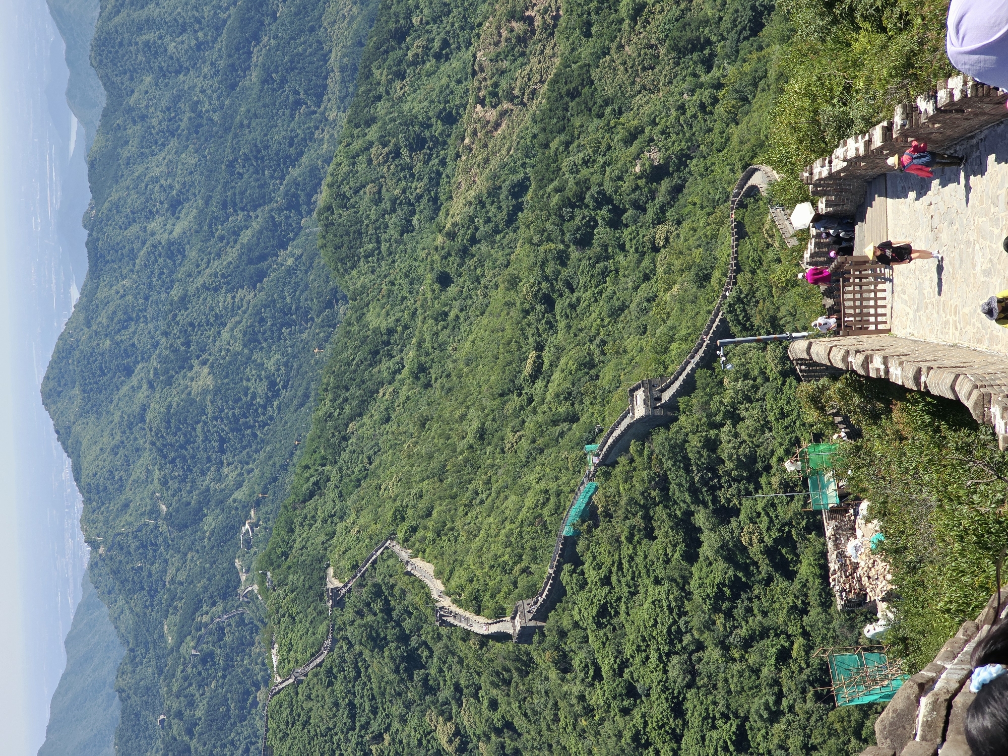 The image depicts a section of the Great Wall of China winding through a lush, mountainous landscape in Huairou District, China. The wall, constructed of stone and brick, is clearly visible, tracing the contours of the rolling green hills. Numerous tourists are present on the wall, walking along its path and descending a stone staircase in the foreground. Some are wearing hats and casual clothing, suggesting a warm, sunny day. The environment is dominated by dense green foliage covering the mountains, indicating a natural, possibly forested, setting. In the middle ground, there are some construction or repair activities underway, with scaffolding and green netting visible. The distant background shows a hazy expanse of sky and possibly a cityscape or more mountains, suggesting a vast, sprawling landscape. The perspective is from an elevated viewpoint, looking down upon the wall and the surrounding terrain.