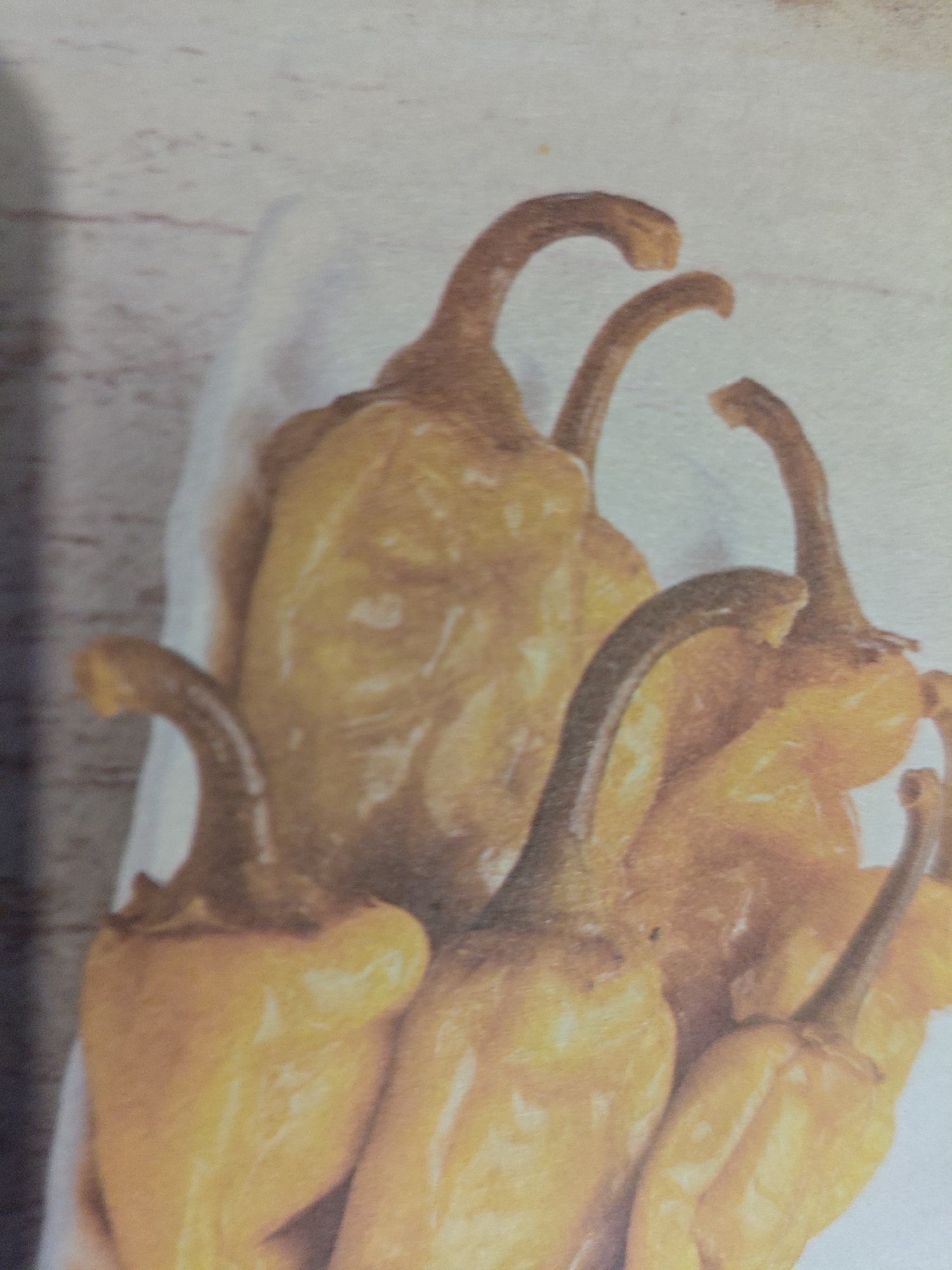 A close-up, slightly out-of-focus, overhead shot shows a cluster of approximately five yellow peppers. The peppers are of the pointed variety and have a glossy sheen. They appear to be arranged on a white surface, possibly paper, which is itself placed on a textured, light-colored background, resembling wood grain. The lighting casts subtle shadows, highlighting the peppers' contours. The image does not provide any specific details related to the city of Armavir, Russia.