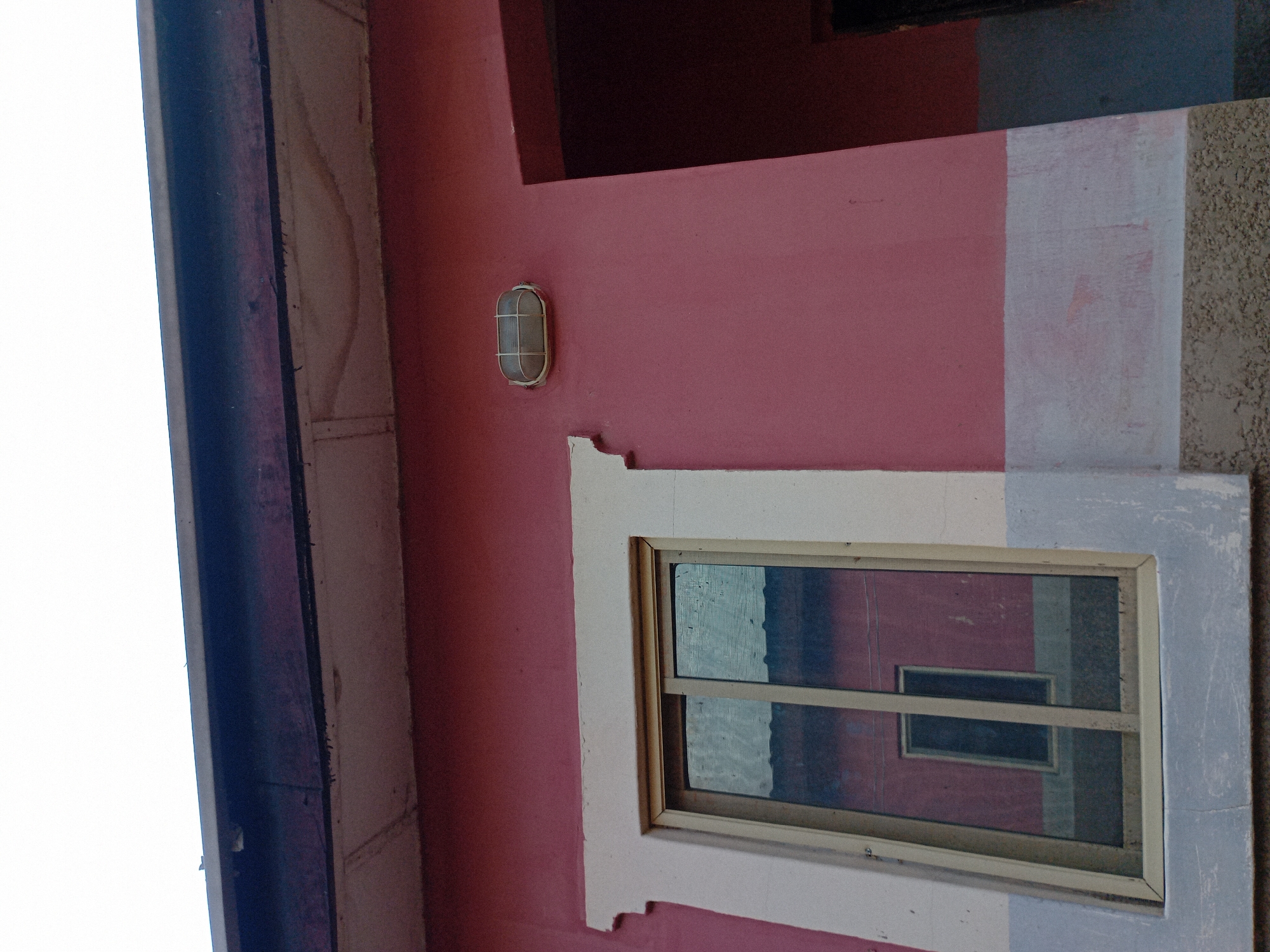 The image shows the exterior of a building in Jauro Yinu, Nigeria, under a bright sky. A pink-painted wall dominates the scene, with a white-trimmed window reflecting another part of the building. Above the pink wall, a dark wooden beam supports a lighter colored soffit. An oval-shaped light fixture is mounted on the pink wall to the right of the window. To the right of the window, a dark opening, possibly a doorway, is visible. The lower part of the building features a horizontal band of lighter paint, followed by a textured, light brown base. The overall impression is one of a simple, functional building in a residential or commercial area of Jauro Yinu.
