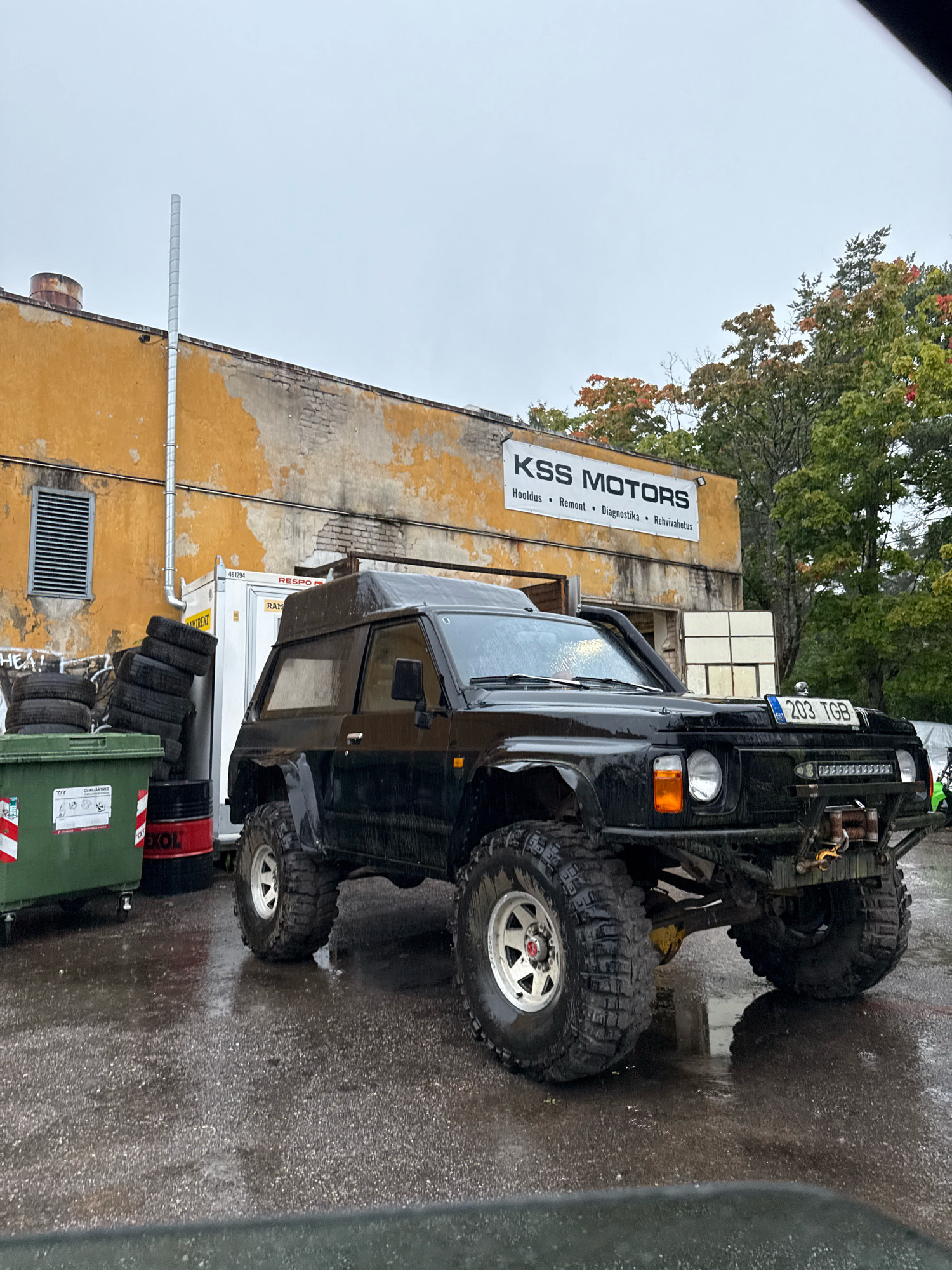 The image depicts an outdoor scene in Tallinn, Estonia, on a rainy day. In the foreground, a black, modified off-road vehicle with large, aggressive tires is parked on a wet asphalt surface. The vehicle has a winch mounted on its front bumper and a light bar. Behind the vehicle is a faded yellow building with a sign that reads "KSS MOTORS" and lists services such as "Hooldus", "Remont", "Diagnostika", and "Rehvihooldus". To the left of the building, there is a green dumpster and a stack of tires next to a black barrel. The sky is overcast, and the overall atmosphere suggests a utilitarian or industrial setting. No people are visible in the image.