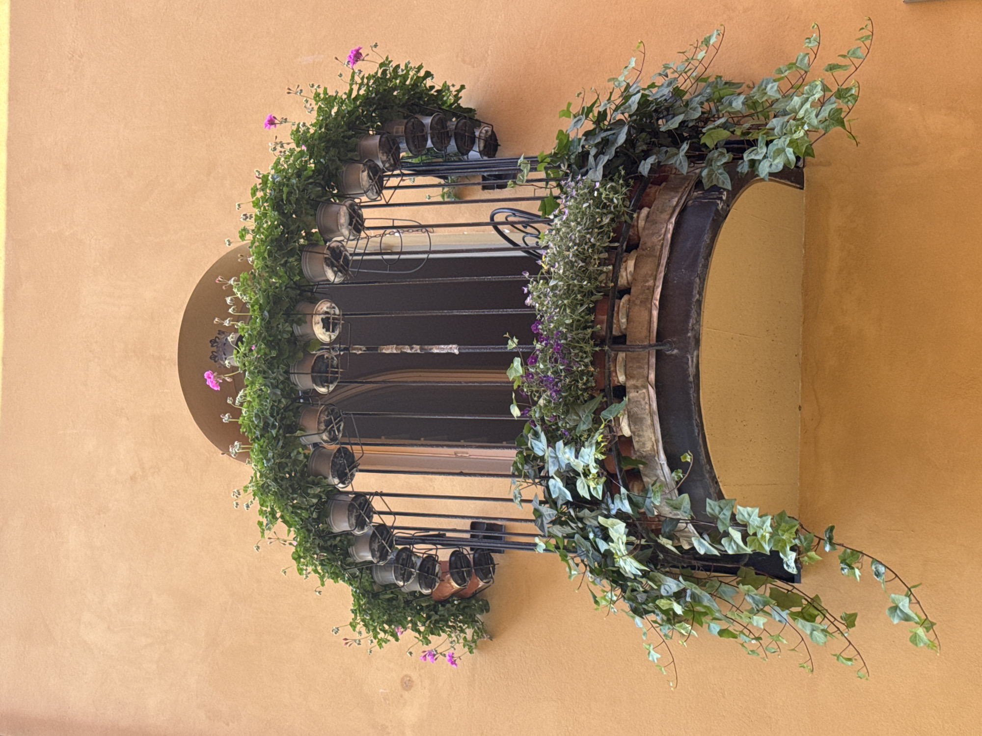 The image shows a balcony overflowing with plants and flowers against an orange stucco wall. The balcony has a dark metal railing and a curved, enclosed bottom. There are numerous potted plants arranged along the top and front of the balcony, with green vines cascading down the sides and front. Some of the plants have small pink flowers. The balcony is situated in front of an arched window with dark bars. The overall scene is one of abundant greenery and floral display. There is no indication that this location is in Stockholm, Sweden.