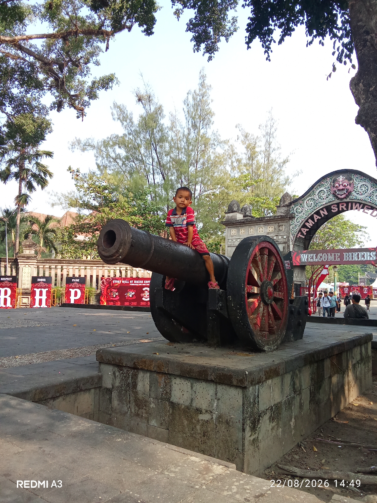 A young boy in a red and blue striped shirt and shorts sits atop a large, dark cannon at the entrance to Taman Sriwedari in Surakarta, Indonesia. The cannon, with red-spoked wheels, is positioned on a stone pedestal. Behind the cannon, an archway displays the text "TAMAN SRIWEDARI" and "WELCOME BIKERS" in large, prominent letters. People are visible walking through the archway. The area is paved, with some vegetation and trees visible in the background. The date stamp on the image reads "22/08/2026 14:49".