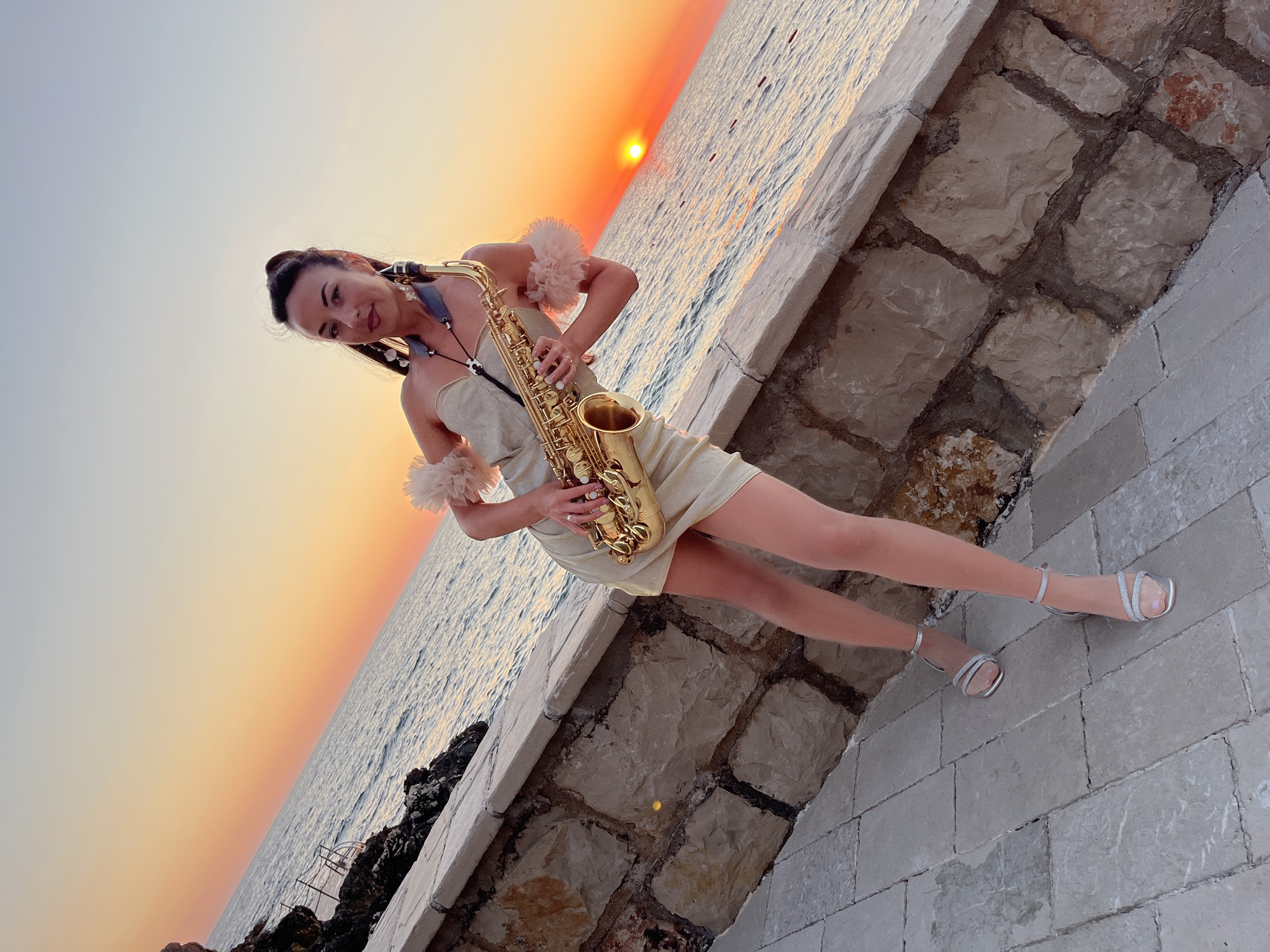 A full-body shot captures a woman standing on a paved walkway next to a stone wall with the sea and a sunset in the background. The woman, who appears to be in her late 20s or early 30s, is wearing a shimmering, light-colored, strapless dress with fluffy accents on the shoulders and silver, strappy high-heeled sandals. She is holding a golden saxophone, with the neck strap around her neck. The sun is setting over the water, casting a warm orange glow. The location is the waterfront in Pečurice, Montenegro, as indicated by the distinct stone wall construction typical of the area and the mountainous coastline characteristic of the Adriatic Sea.