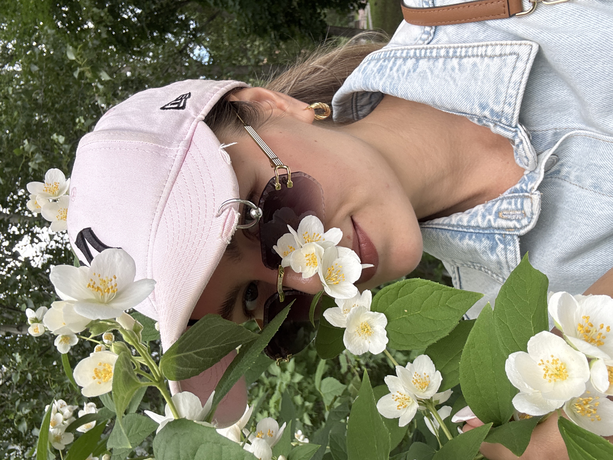 A young woman with blonde hair and fair skin is partially obscured by blooming white jasmine flowers. She is wearing a light pink baseball cap, dark-lensed sunglasses with gold frames, and a light blue denim jacket. The woman has a gold hoop earring in her right ear. She is outdoors, with lush green foliage and trees visible in the background. The scene suggests a casual, possibly daytime setting in a park or garden.