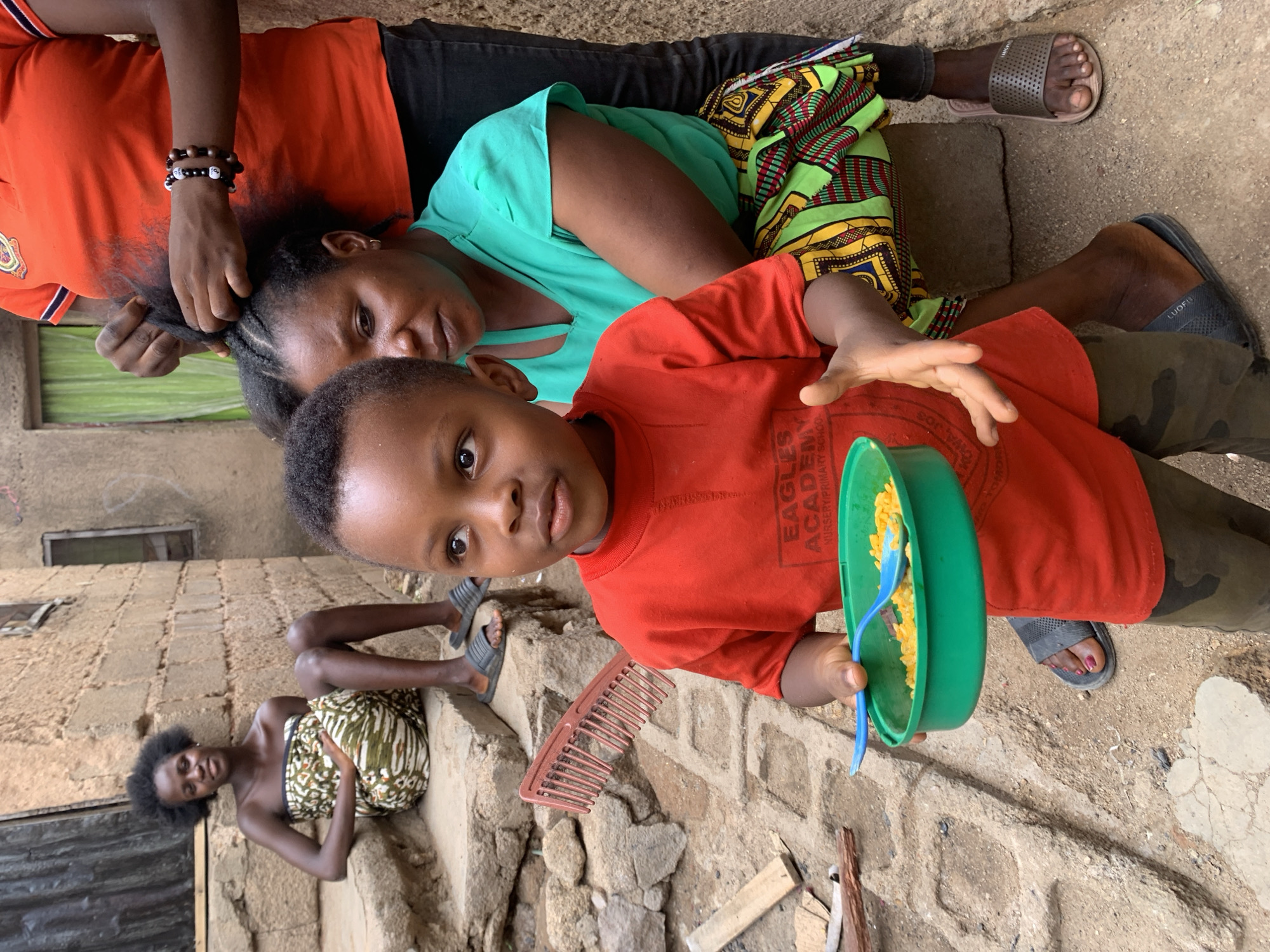 Jos, Nigeria: An outdoor scene depicting three women and a boy, potentially in a courtyard or an area adjacent to a building with stone or concrete walls. One woman is seated on the ground wearing a green top and a patterned skirt while someone with an orange top braids her hair. A young boy in an orange shirt and camouflage pants stands in front of them, holding a green bowl with a blue spoon. A broken comb rests on the ground next to him. In the background, another woman is sitting on a step or ledge, wearing a patterned wrap. The environment is earthy and somewhat worn.