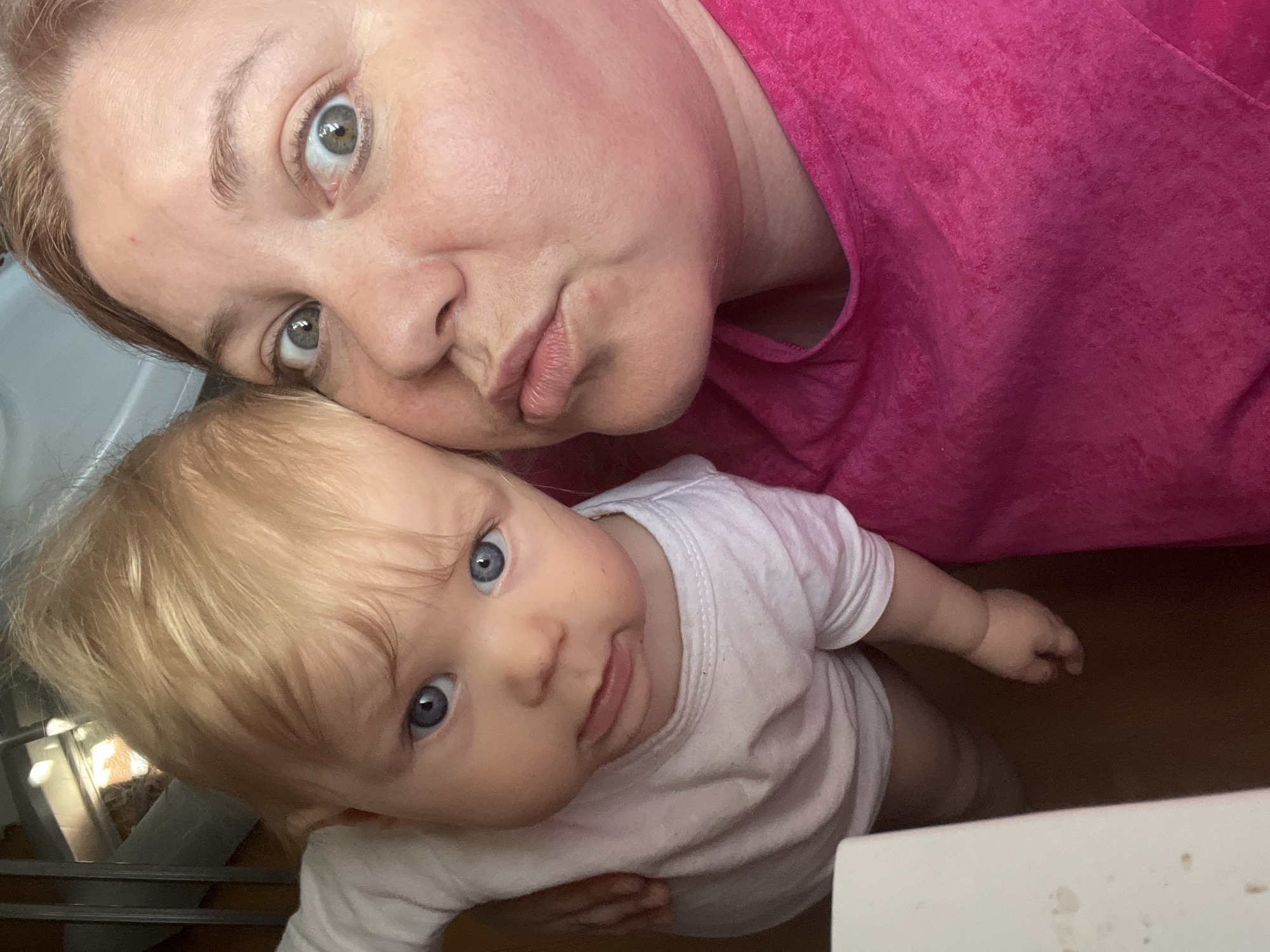 A selfie featuring a woman and a baby. The woman, fair-skinned with red hair, is wearing a pink top and puckering her lips at the camera. The baby, fair-skinned with blonde hair and blue eyes, is wearing a white shirt and looking up at the camera. The background is out of focus, but appears to be a room with a white wall and a crib. No location information in Vesyoloye, Russia can be determined from the provided image.