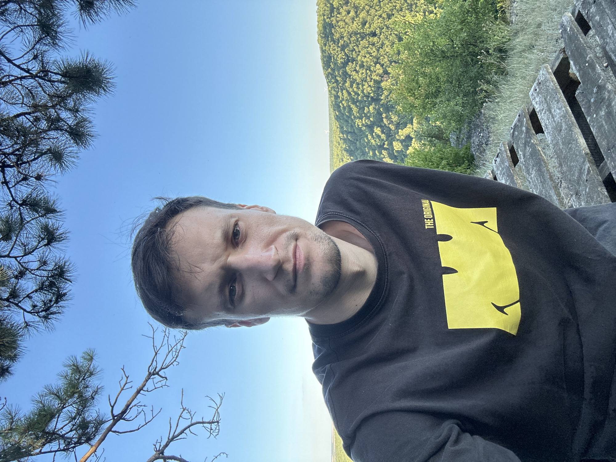 A man takes a selfie outdoors in the city of Pryvorottia Druhe, Ukraine. The man is wearing a black t-shirt with a yellow graphic. He is in the foreground, with a lush, green forest and a clear blue sky in the background. There are also some trees branches at the top of the image. The man is looking directly at the camera. A wooden pallet is visible on the right side of the image.