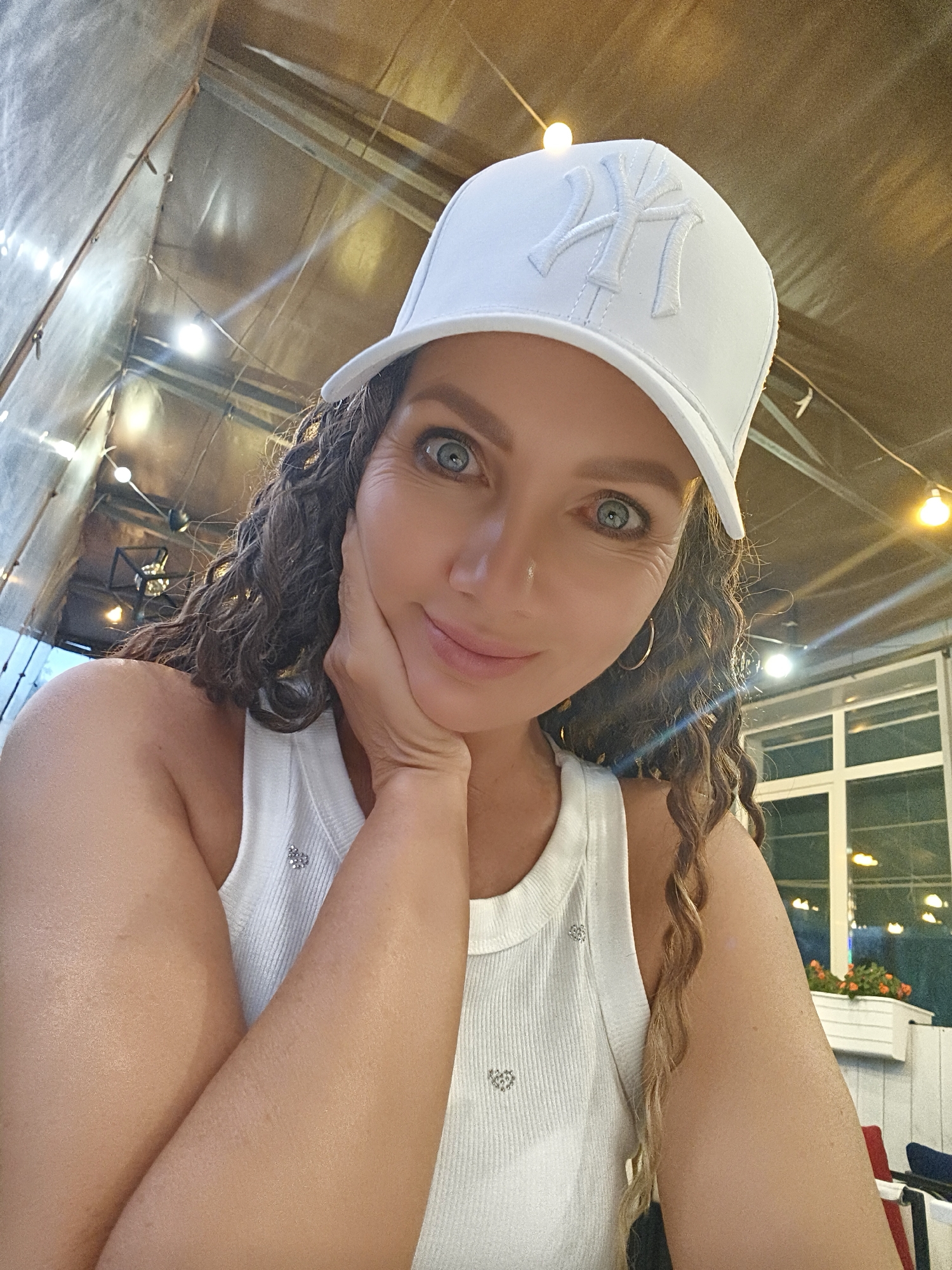 The image is a selfie of a woman wearing a white baseball cap with a white "NY" logo. She has curly brown hair, blue eyes, and is resting her chin on her hand. She is wearing a white tank top with small heart-shaped embellishments. The background appears to be an outdoor seating area with exposed beams and string lights, suggesting it might be evening. Through a window in the background, potted flowers can be seen. The lighting is warm and ambient. The woman's expression is relaxed and engaging.