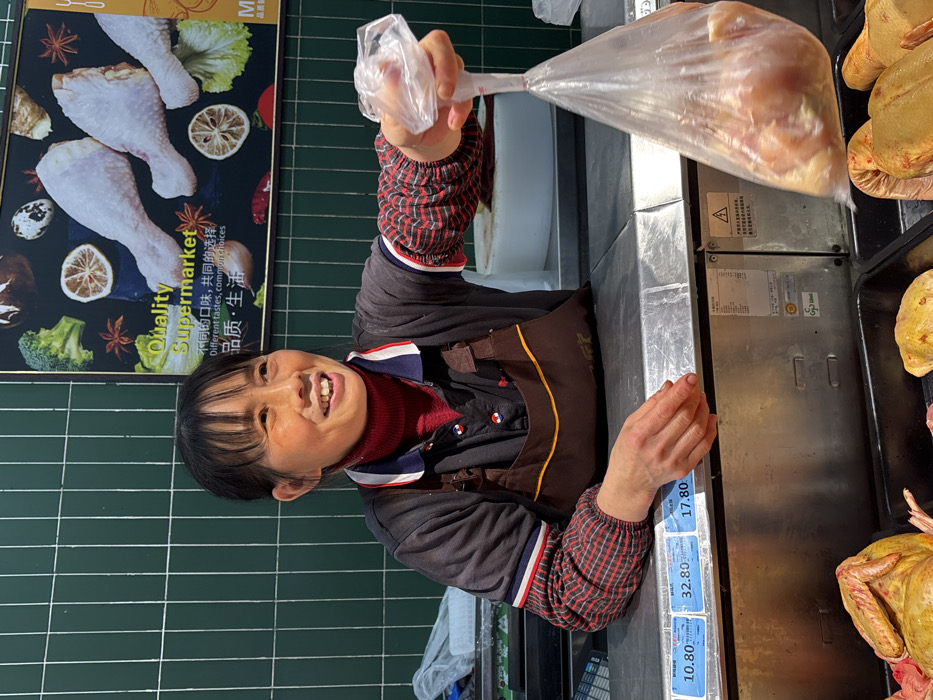 The scene is set in a fresh food section of a supermarket in Jiangjin, China. An adult female vendor, with dark hair and bangs, stands behind a stainless steel counter, smiling and looking towards the right side of the frame. She wears a dark long-sleeved shirt over a red turtleneck, with a brown apron, and a red and black plaid undershirt visible on her left arm. Her right hand is extended forward, holding a transparent plastic bag containing raw poultry pieces. Her left hand rests on the metal counter. To her left on the counter, a digital display shows numeric values: "10.80," "32.80," and "17.80," likely indicating prices or weights. In the immediate foreground, below the counter level, raw whole poultry items, possibly chickens, are displayed in dark trays. The background consists of a vertical green tiled wall and a large promotional poster for "Quality Supermarket," which features images of various food items including raw chicken drumsticks, broccoli, mushrooms, lemons, and spices, alongside text in both English and Chinese. Behind the vendor, on the counter, a white circular cutting board and stainless steel utility units with Chinese labeling are visible.