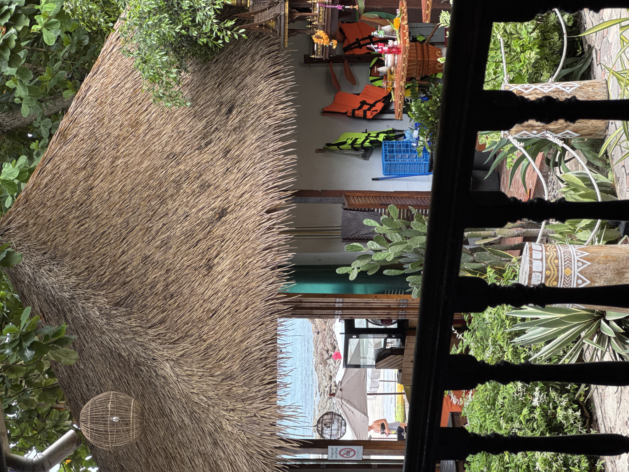 This image depicts an outdoor scene at a location in Ao Phai, Thailand, possibly a beachside establishment. In the foreground, a dark wooden railing with turned balusters obscures part of the view. Beyond the railing, lush green foliage and various plants, including a cactus and an agave plant, are visible. A weathered wooden post with a painted geometric design stands amongst the greenery.
The midground reveals a thatched-roof structure, characteristic of tropical architecture. Underneath the eaves of this structure, several orange life jackets and a bright green life jacket are hanging. A blue plastic basket is also present. A small, ornate shrine with offerings, including marigolds and bottles, is situated on a table. A glass cabinet or counter is visible to the left, with what appears to be a refrigerator behind it. A person wearing a bikini top is standing near a beach umbrella on the sandy beach in the background. The sea, with calm water and rocky shore, stretches to the horizon under an overcast sky. A woven pendant light hangs from the thatched roof.