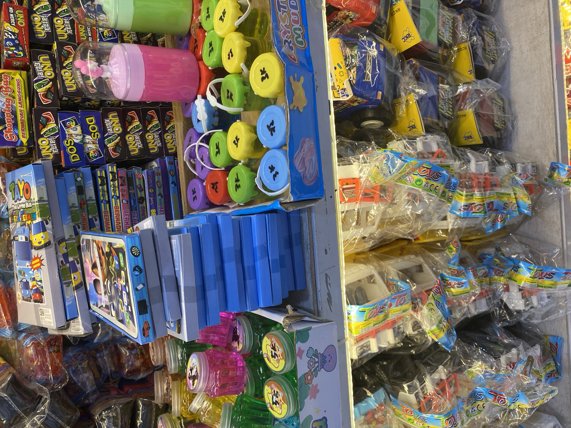 The image depicts a retail display of toys and novelty items in what appears to be a small shop or market stall. The items are predominantly colorful, with a focus on children's products.
In the foreground and mid-ground, there are various types of toys. These include small jars of colored slime or putty, some with cartoonish characters on the lids. There are also stacks of boxes, some appearing to be card games like UNO, and other boxed toys with cartoon imagery. A prominent pink plastic container with a clear lid, containing a pink toy figurine, sits on a display. Next to it is a green container, also with a clear lid. There are also many small, colorful buckets with lids, some containing a gel-like substance.
The background shows more stacked merchandise, including more boxes of games and other packaged goods. Some of these packages are transparent, revealing toy vehicles or other small items within. The overall impression is one of a busy, somewhat cluttered retail environment focused on selling a wide variety of inexpensive toys.
The context provided suggests this scene is located in Lamteumen Barat, Indonesia. While the image itself does not contain explicit geographical markers, the merchandise and the style of display are consistent with the types of goods often found in local markets and small shops in such regions. There are no people visible in the image. The lighting suggests an indoor environment, possibly with artificial light sources. The surfaces appear to be made of wood or particle board for the shelves.