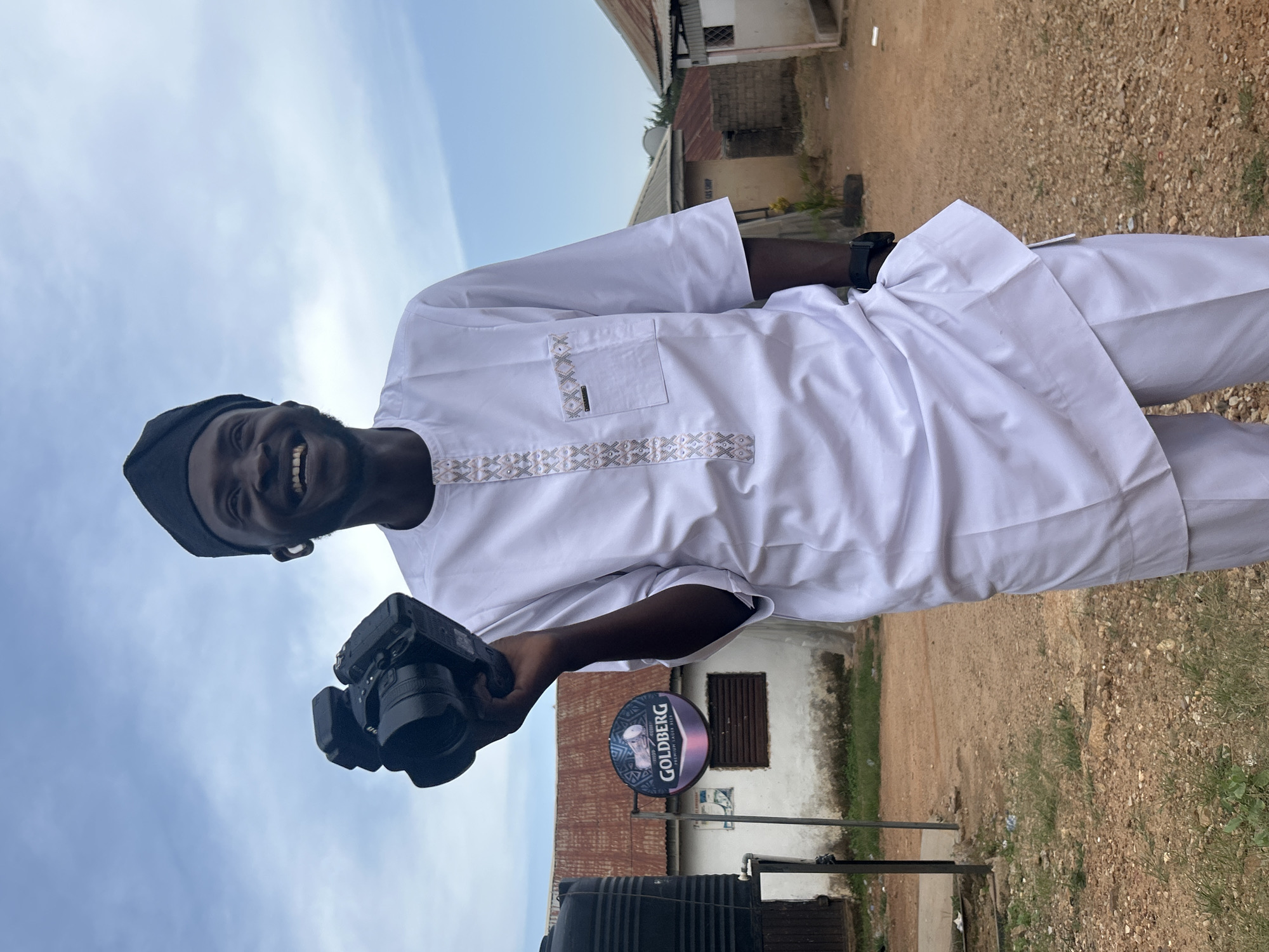 A man smiles at the camera while holding a DSLR. The man is wearing a white garment with embroidered detail around the collar and pocket and a black kufi cap. A "Goldberg" beer sign is behind the man. The ground is made up of dirt and stones. Some buildings can be seen in the background. The photograph appears to have been taken in Jos, Nigeria.