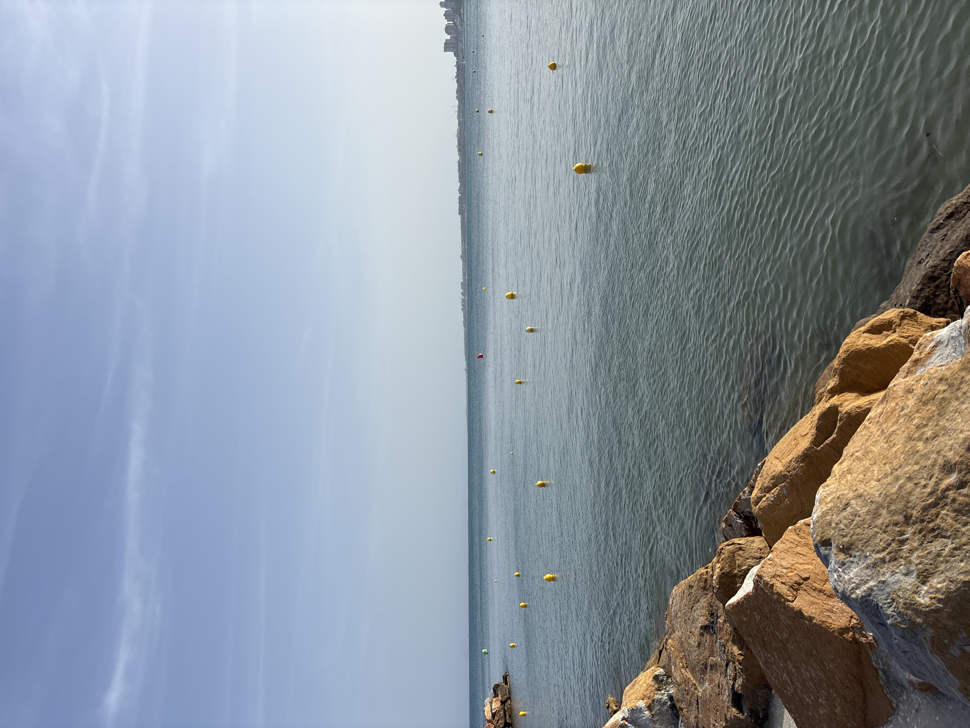 The image depicts a view of the Mediterranean Sea from the coast of Orihuela, Spain. In the foreground, large, weathered rocks form a natural barrier along the shoreline. The sea stretches out to the horizon, with numerous yellow buoys floating on its surface, indicating a designated swimming or boating area. The water is calm with gentle ripples. In the distance, a faint outline of buildings and structures can be seen along the coastline, suggesting an urban or developed area. The sky is clear and blue with a few wispy clouds. The overall scene is tranquil and captures a typical coastal landscape.