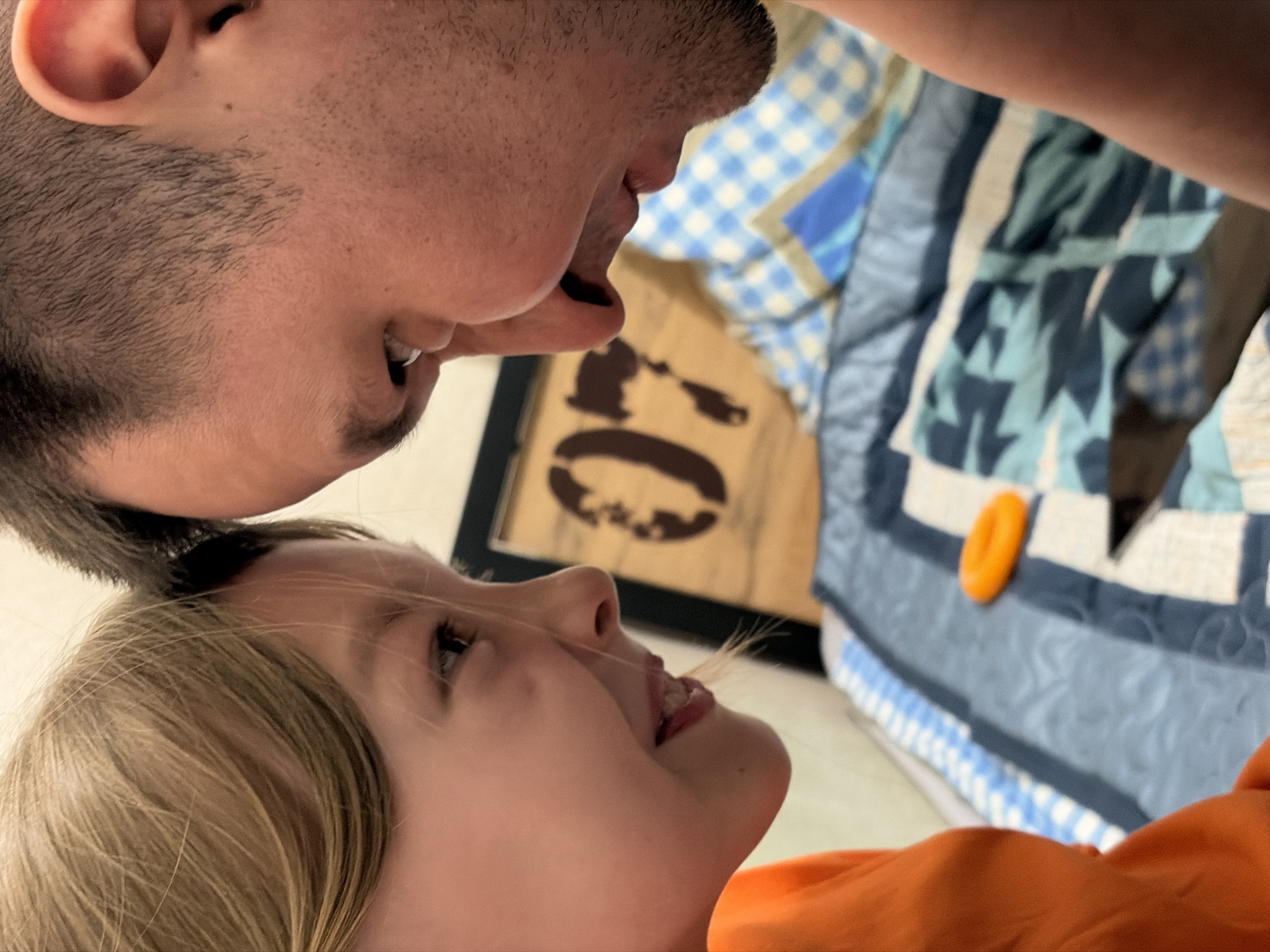 A close-up, tilted shot depicts a man and a young girl, possibly a father and daughter, in close proximity. The man is positioned above the girl, his face partially visible from the nose up, including his mouth slightly open with a glimpse of his teeth. He has short, dark hair and stubble. Below him, the young girl looks up at him with her mouth also slightly open. She has light hair and fair skin.
In the background, out of focus, is a quilt with blue and white patterns, as well as a framed object with a dark design on a light background. An orange plastic toy is also visible on the quilt.
There is no information in the image to determine its location in Izhevsk, Russia or any other location. The scene appears to be indoors.